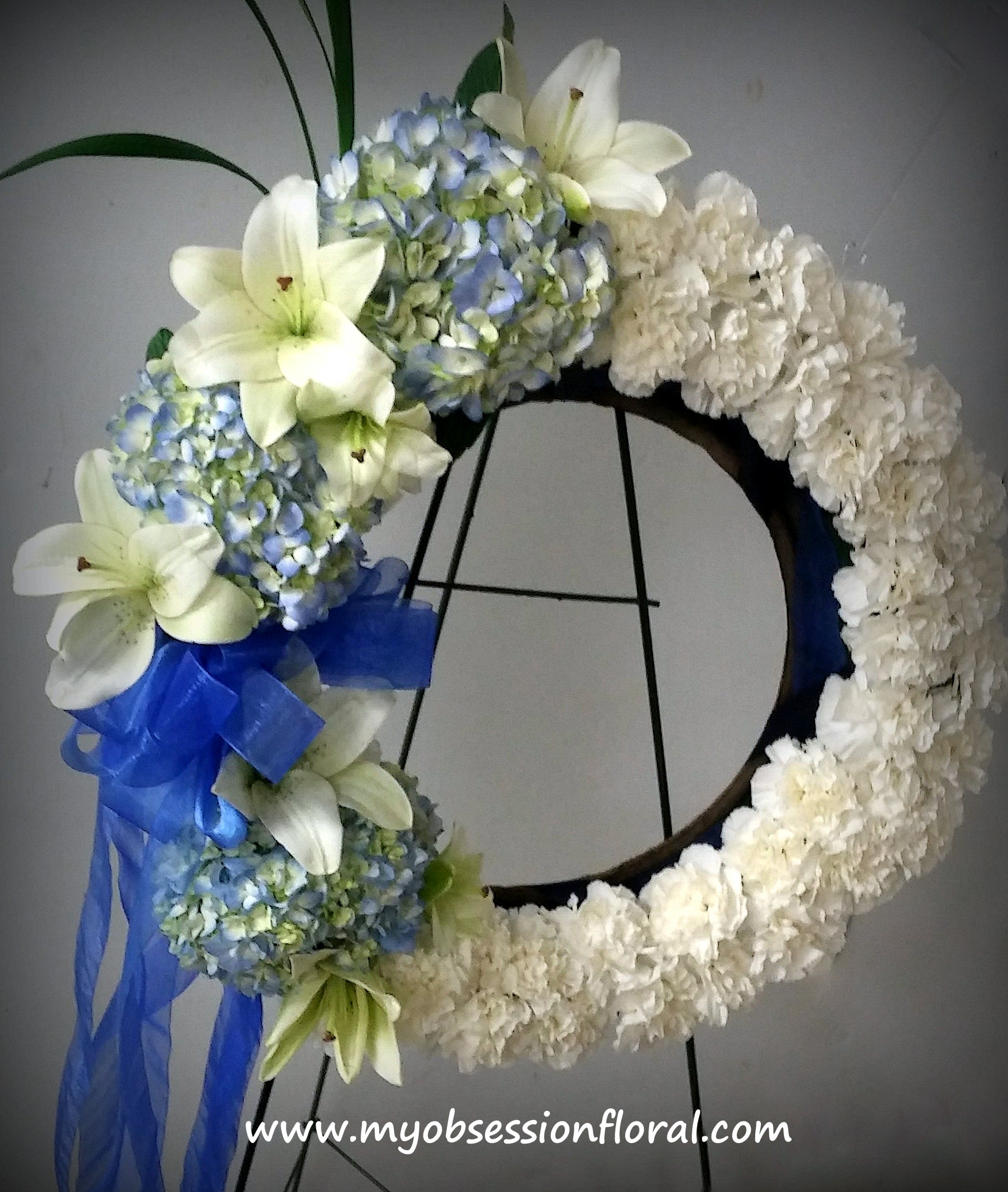 White floral wreath with blue hydrangeas, lilies, and a blue ribbon