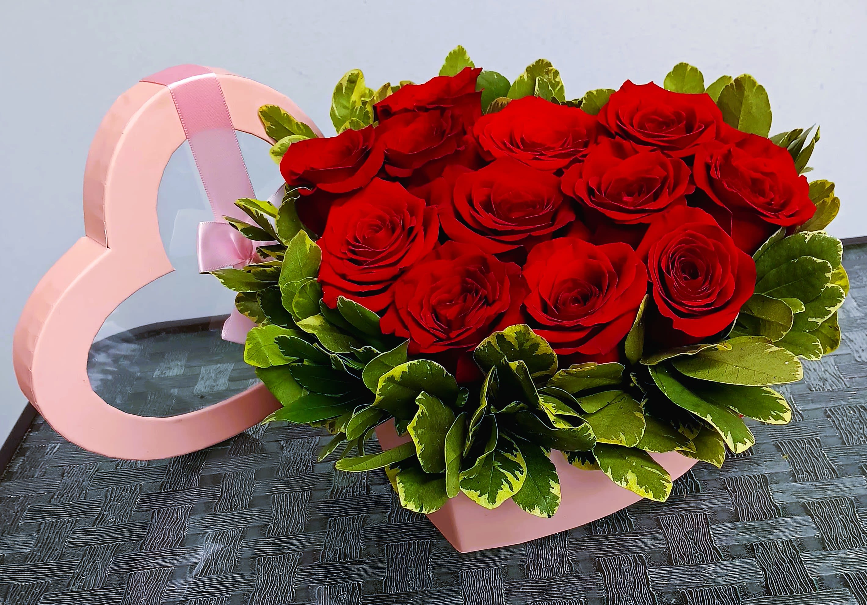 Red roses arranged in a pink heart-shaped box with green foliage