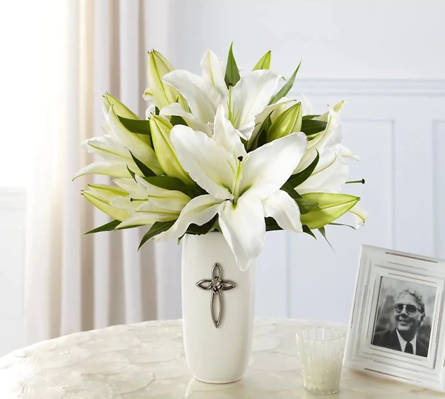 White lilies arranged in a tall white vase with a cross emblem