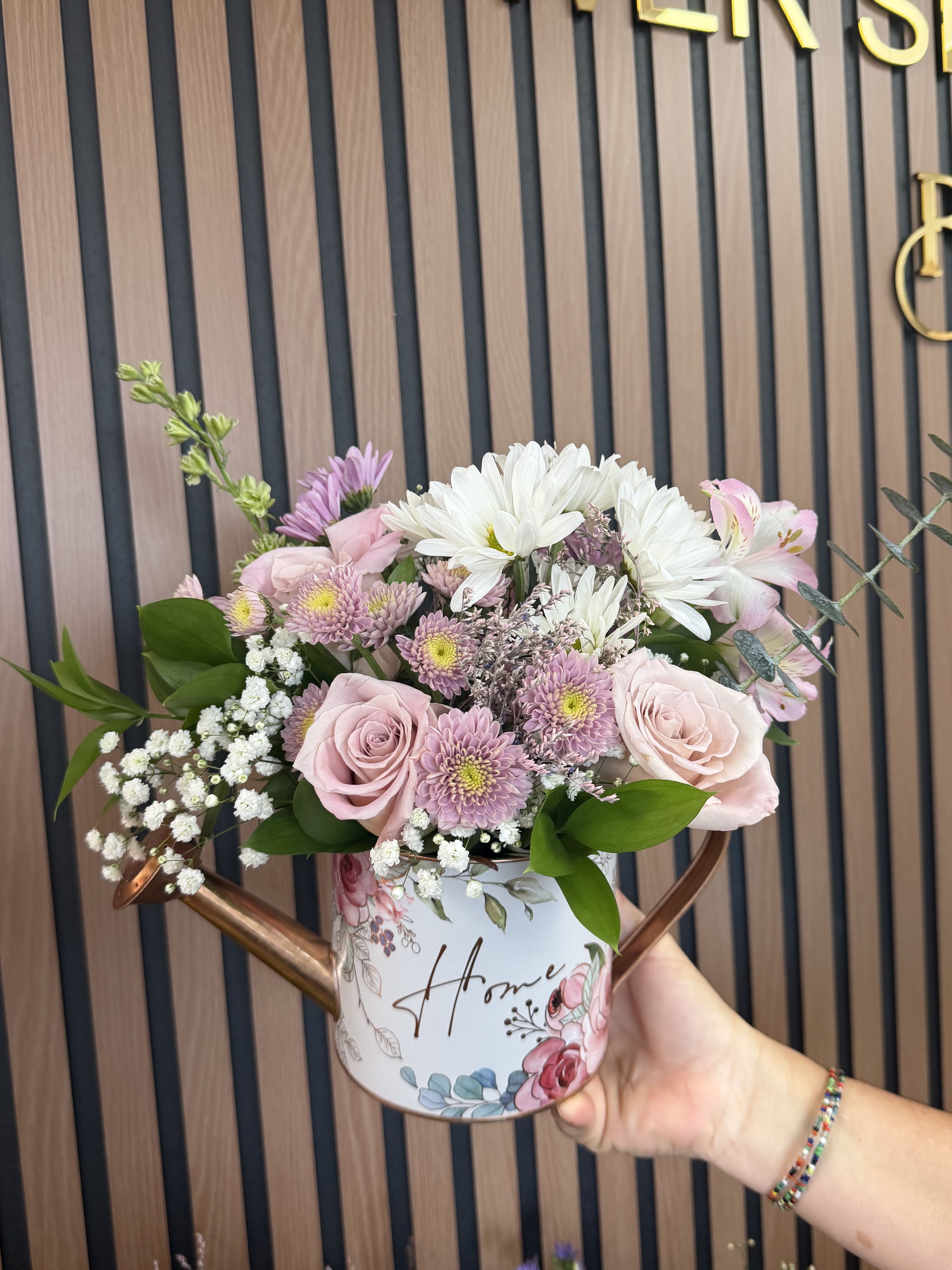 Lovely May - Pale pink roses and soft lavender chrysanthemums spill from a charming decorative watering can to create a gentle, welcoming flower arrangement perfect as a birthday bouquet, housewarming gift, or thoughtful Mother's Day surprise. This flower arrangement for thinking of you moments features white daisy-like blooms, alstroemeria accents and clusters of baby's breath arranged in a rounded, abundant style so the recipient immediately feels seen and celebrated.  The mix of pink, white, green and purple blooms sits above the rim in a floral-printed metal watering can-complete with a copper-toned handle and the word "Home" on the container-so it doubles as décor after the flowers fade. Textures range from glossy green leaves to the soft, cloud-like baby's breath and the velvety roses; overall dimensions present as a full, casual centerpiece sized for tabletops and counters. Ideal for saying thank you, cheering someone up just because, or brightening a new space, this easy-care arrangement arrives ready to display-no extra vase needed.