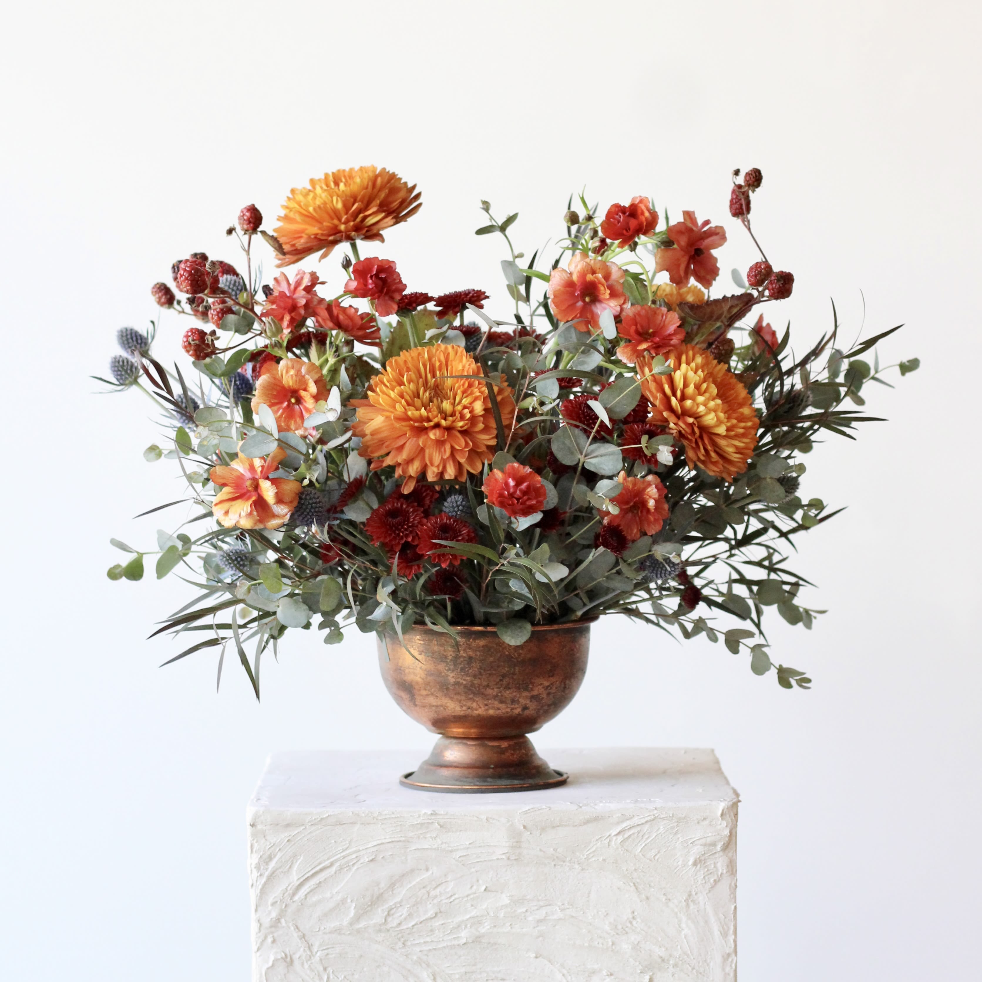 Thanksgiving Collection: Autumn Embers - Introducing Autumn Embers—the perfect centerpiece for your Thanksgiving table. This stunning arrangement features a vibrant mix of golden orange cremone mums, butterfly ranunculus, blue thistle, and fruited blackberry. Its unique jewel-toned palette captures the essence of fall, adding warmth and elegance to any gathering. Housed in a charming copper compote, Autumn Embers not only elevates your decor but also brings a touch of nature's autumnal beauty indoors. Ideal for Thanksgiving hosts looking to impress their guests with a one-of-a-kind floral masterpiece. Celebrate gratitude and the season's splendor with Autumn Embers!