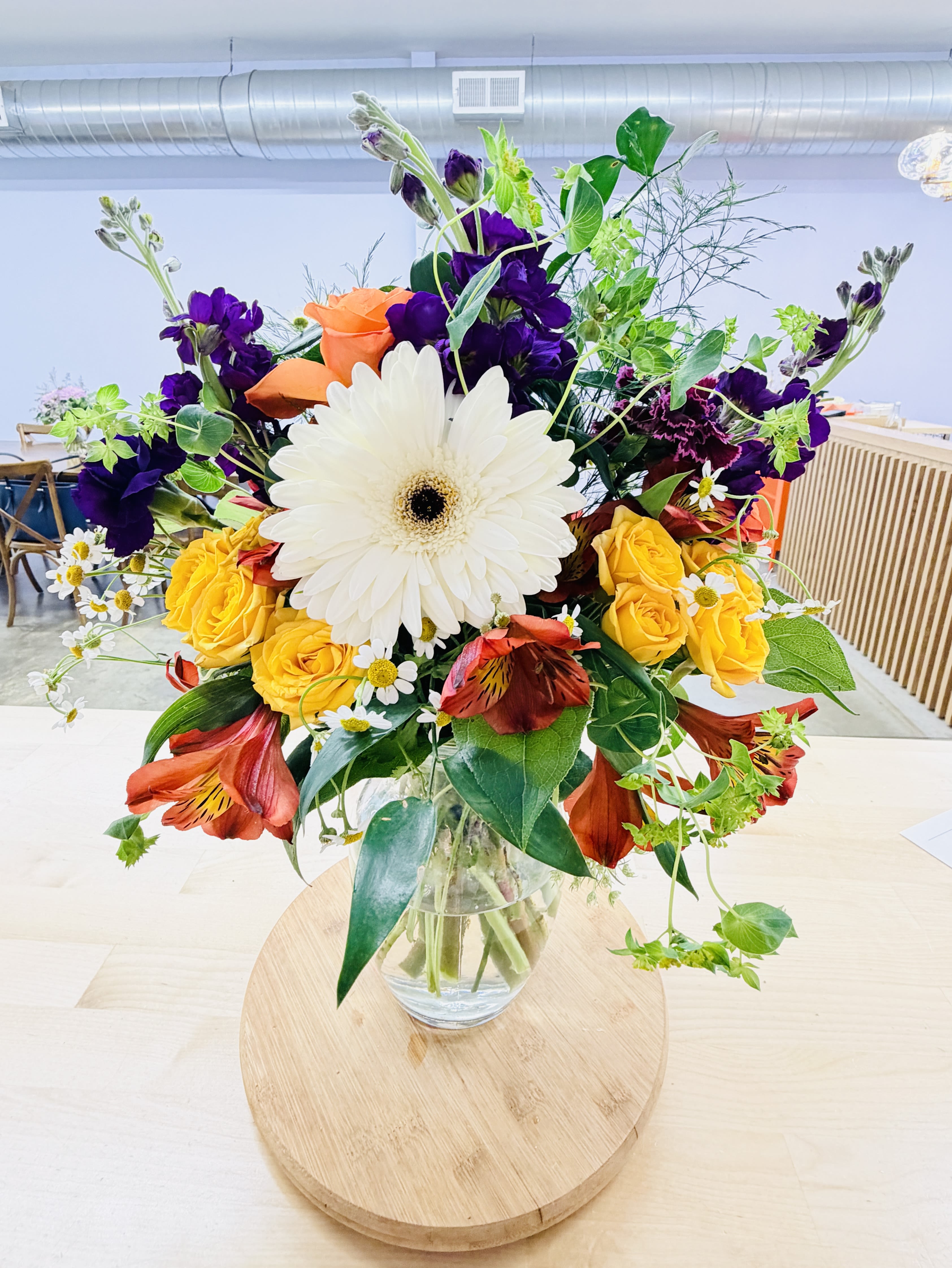 In Love with Daisy (Vibrant) - A bright 'star of the show' gerbera daisy (color may vary) crowns this mixed-color arrangement, creating an immediate focal point that feels joyful and fresh - perfect as a birthday bouquet, thank-you gift, or everyday celebration arrangement. The mix of roses, yellow or orange alstroemeria, stock and small white daisy-like accents gives the recipient a vivid, high-contrast palette that reads as happy and energetic rather than fussy.  Arranged loose and garden-style in a clear glass vase, the bouquet has layered texture from airy greenery and filler stems that lend movement and a slightly wild, abundant look. The clear vase keeps the focus on saturated blossoms and lets stems show through for a natural, modern presentation.