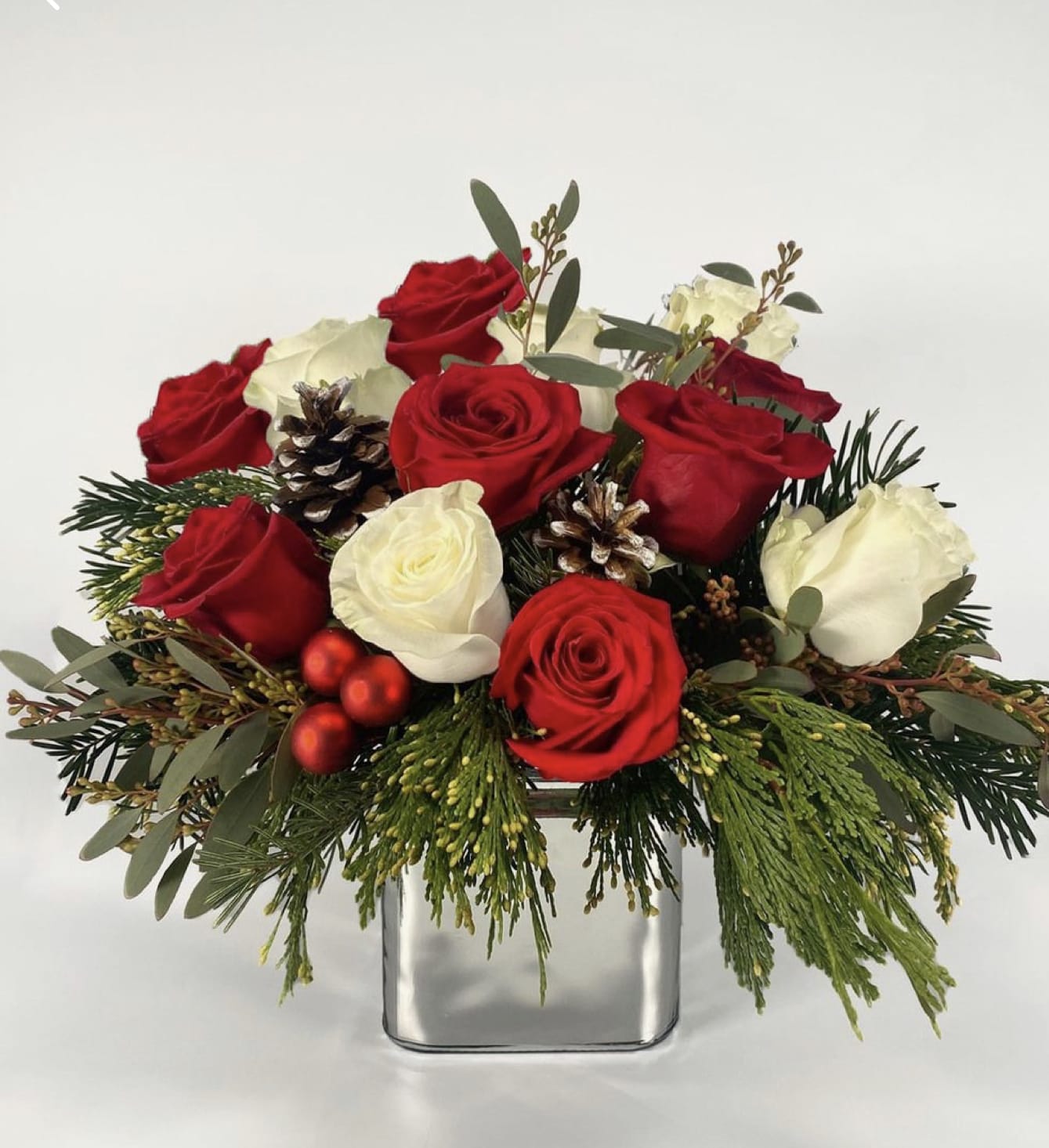 Red and white roses arranged in a square glass vase with pinecones and greenery
