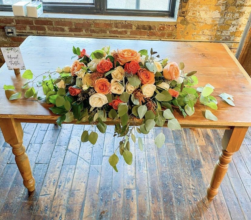 Long low bouquet of orange and cream roses with trailing greenery on a wooden table