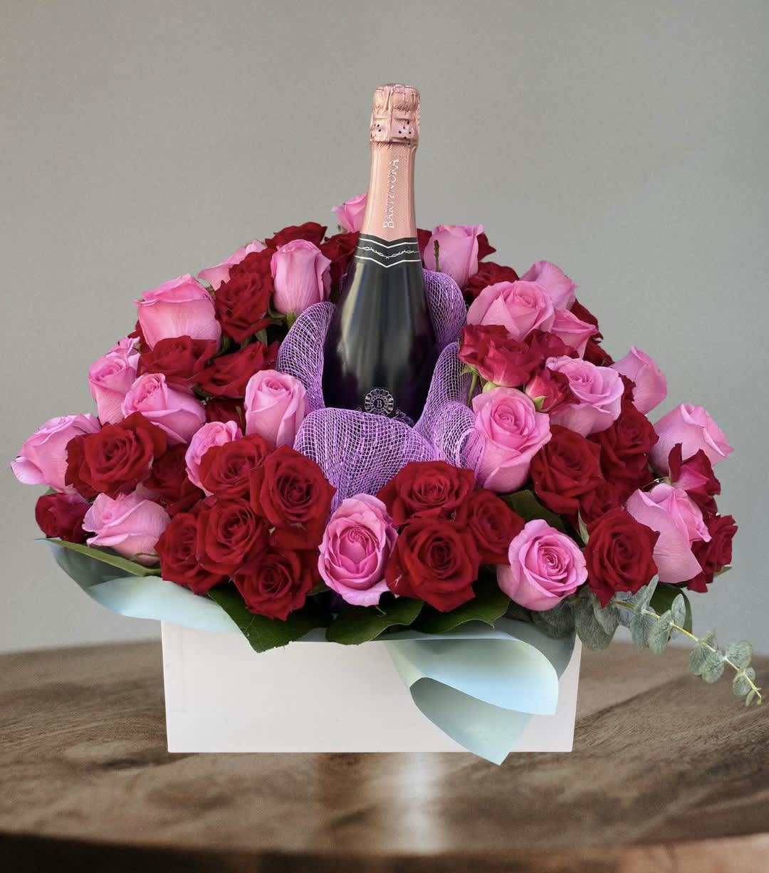Pink and red roses arranged around a champagne bottle in a white box