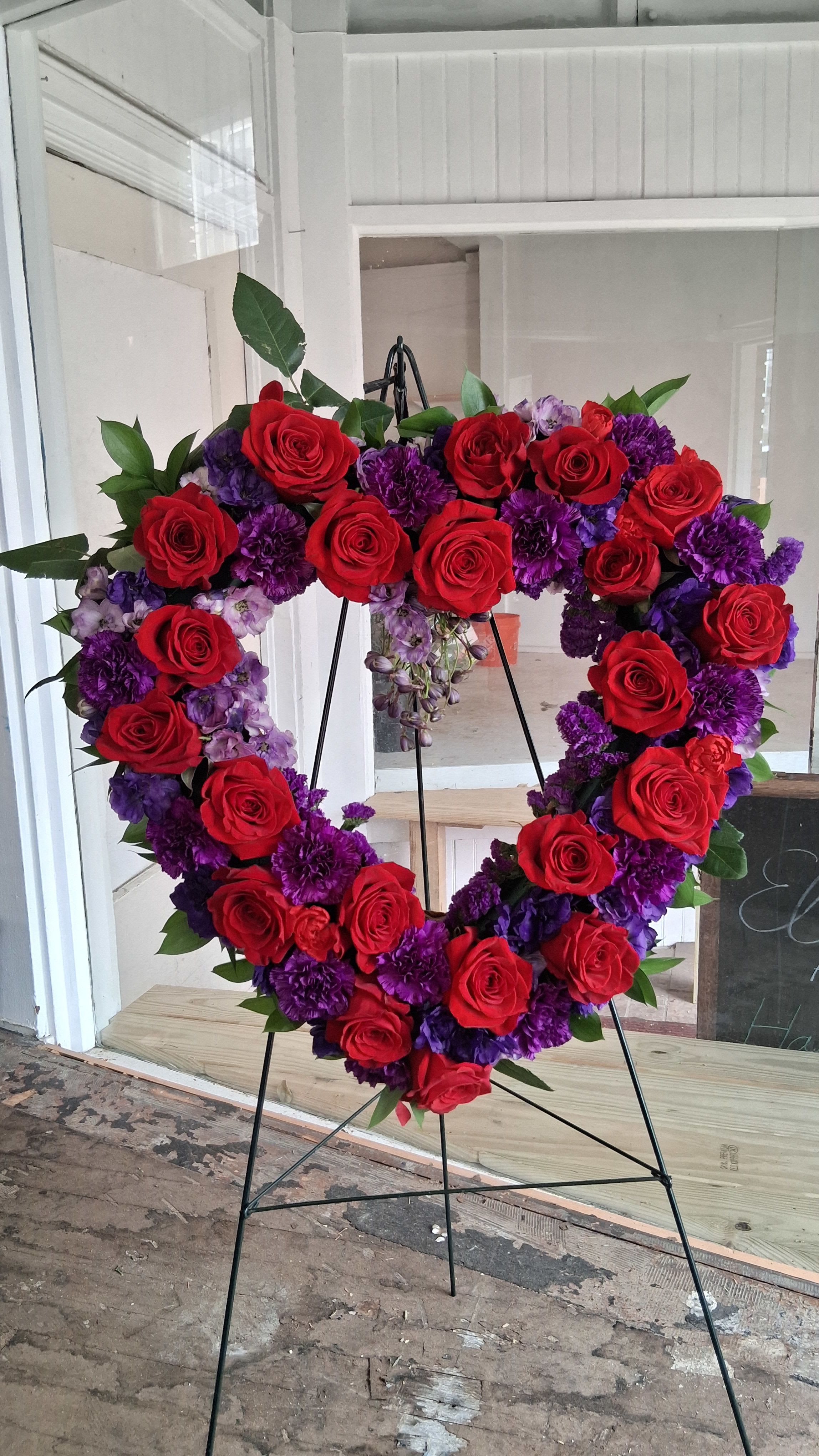 Purple heart - Purple Heart” is a beautifully crafted heart‑shaped wreath featuring a dramatic blend of deep red roses and lush purple blooms. Each flower is placed with intention, creating a rich, dimensional design that symbolizes devotion, remembrance, and heartfelt honor. The vibrant color palette offers a modern yet timeless look, making this wreath a powerful choice for memorials, celebrations of life, or any service where love and respect need to be expressed with beauty and strength.  Hand‑designed in our studio and mounted securely on a standing easel for a graceful presentation.