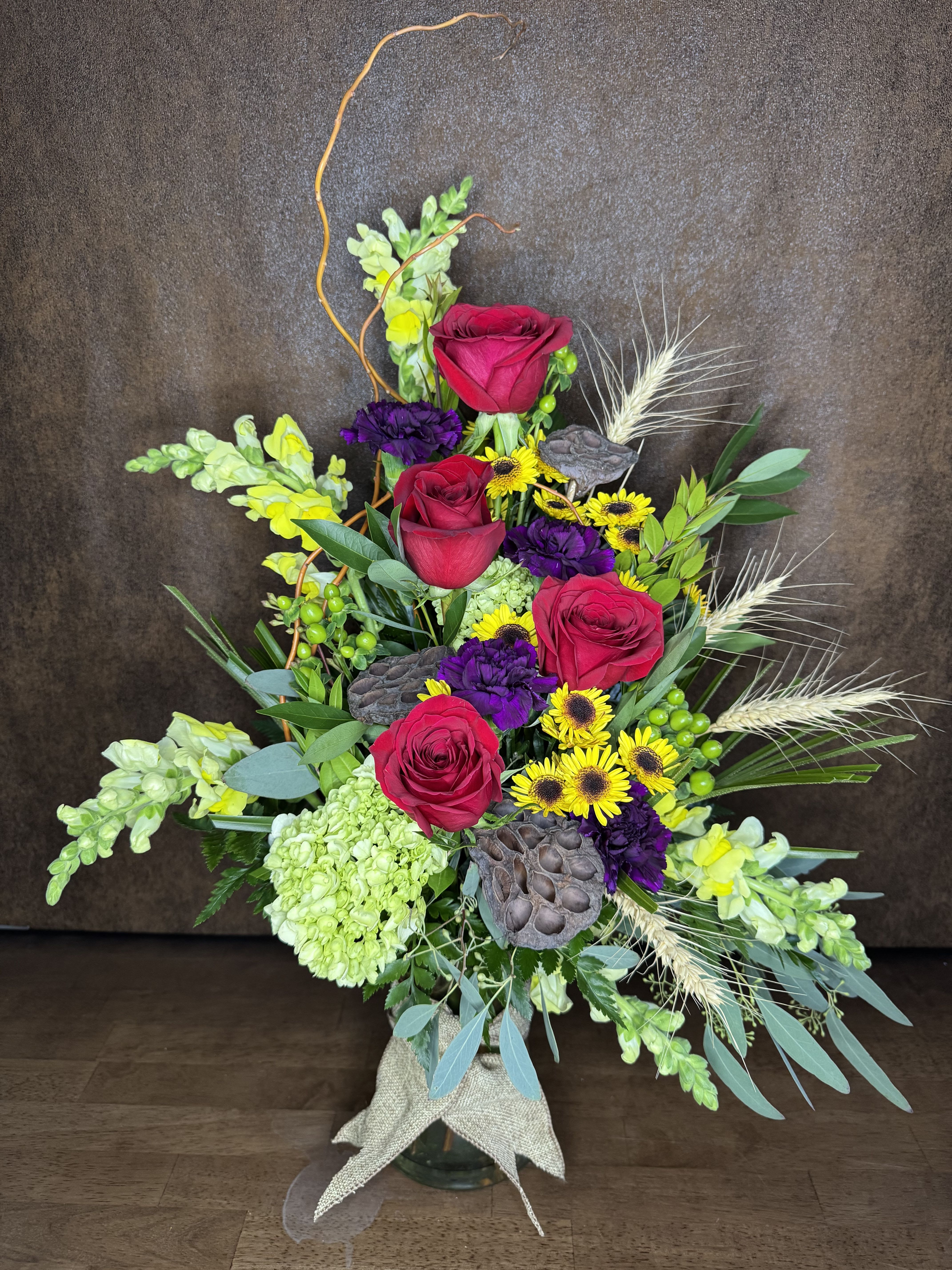 Tall mixed bouquet with red roses, yellow daisies, and purple blooms in a glass vase