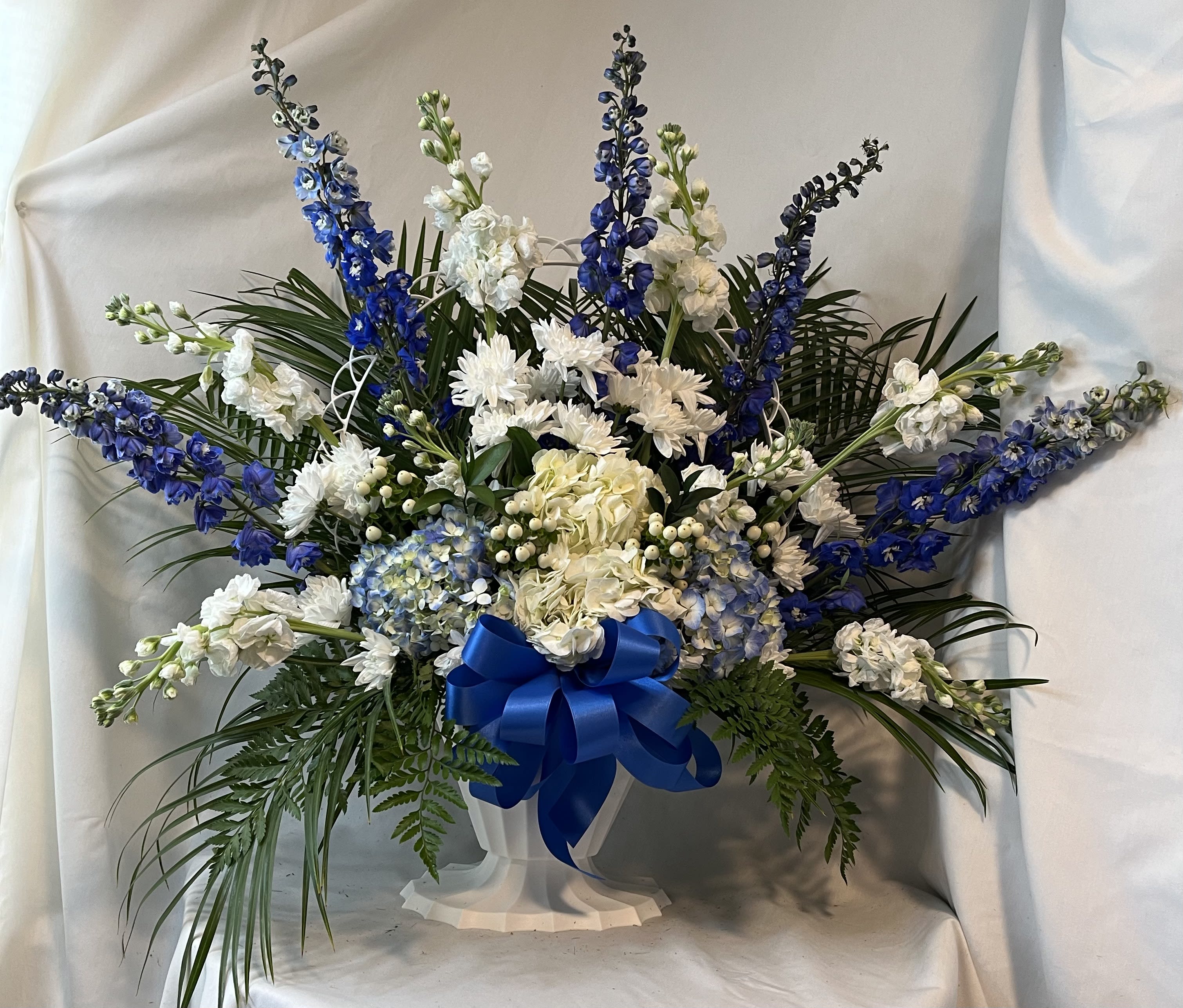 Blue and white floral arrangement in a white pedestal vase with a blue ribbon