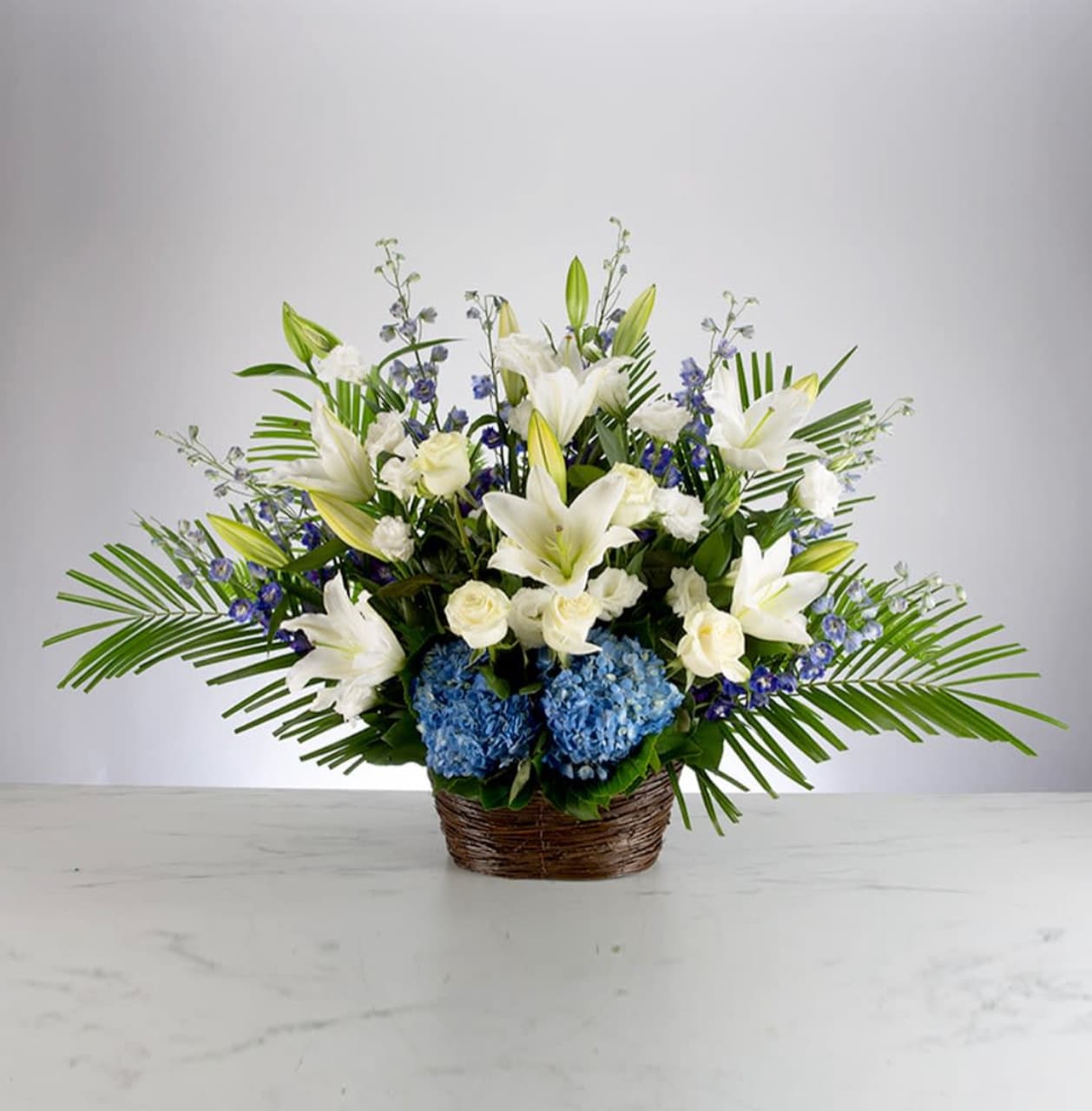 Deepest Condolences - A blue, white, and green funeral basket for any type of service. Held in a wicker basket, large palms spread out and give a natural spotlight for the feature blooms and colors.