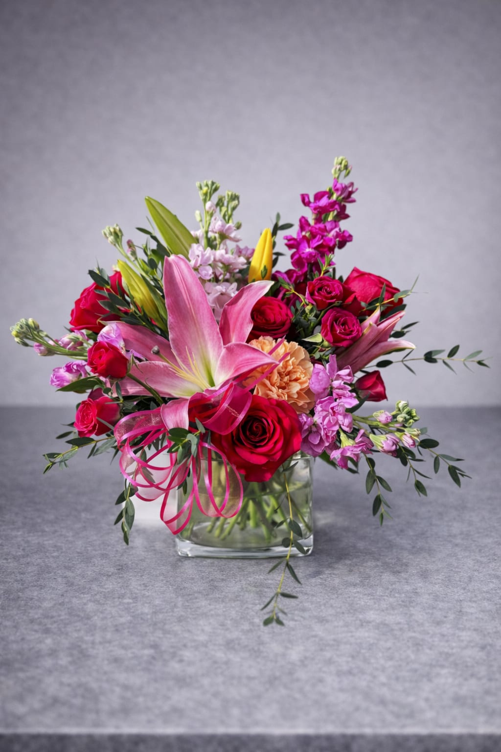 Pink lilies and red roses arranged in a clear glass vase with ribbon.