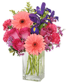 Pink gerberas, roses, carnations, and purple irises in a clear glass vase