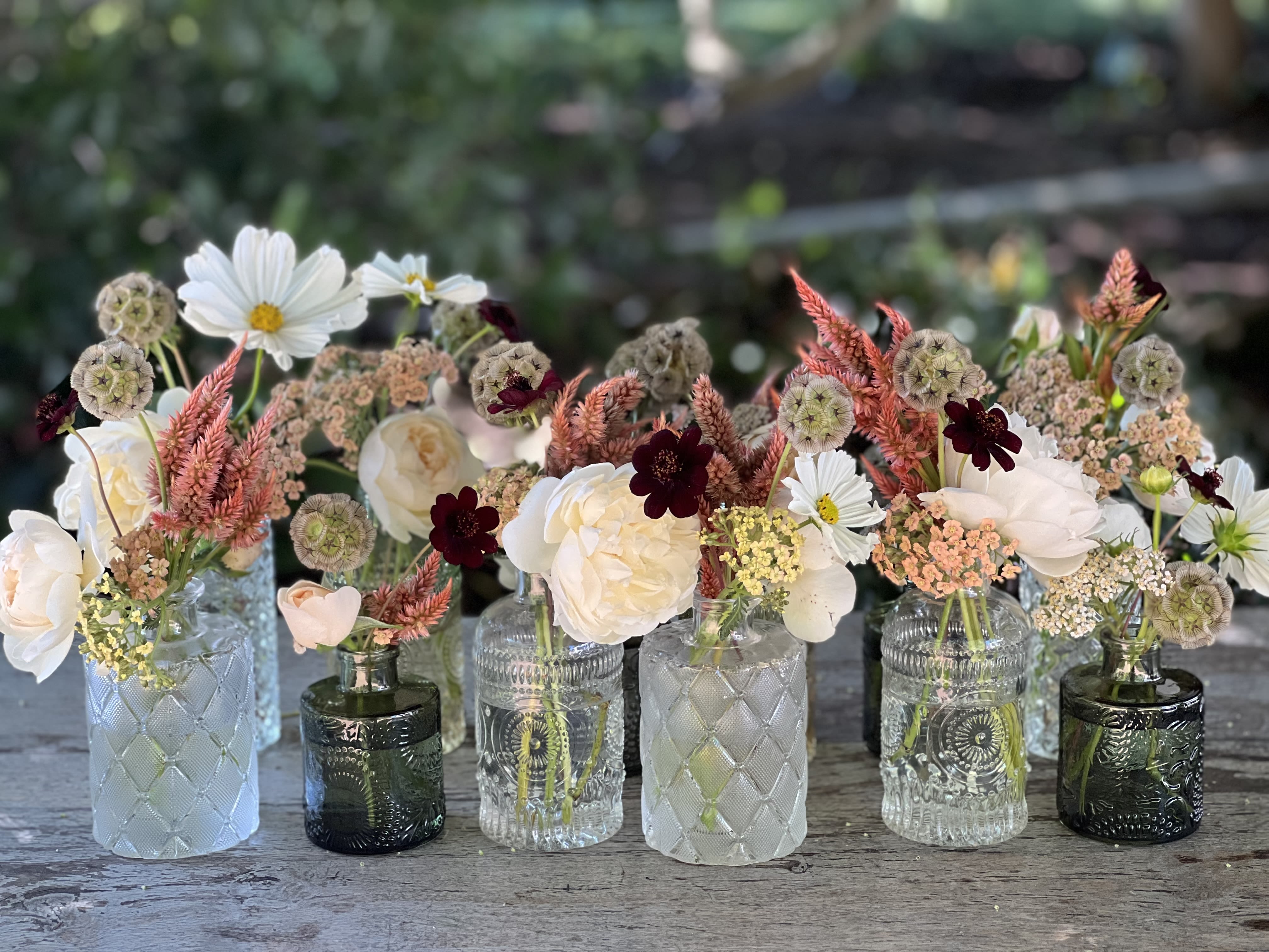 Small mixed flower arrangements in assorted glass bottles on a table
