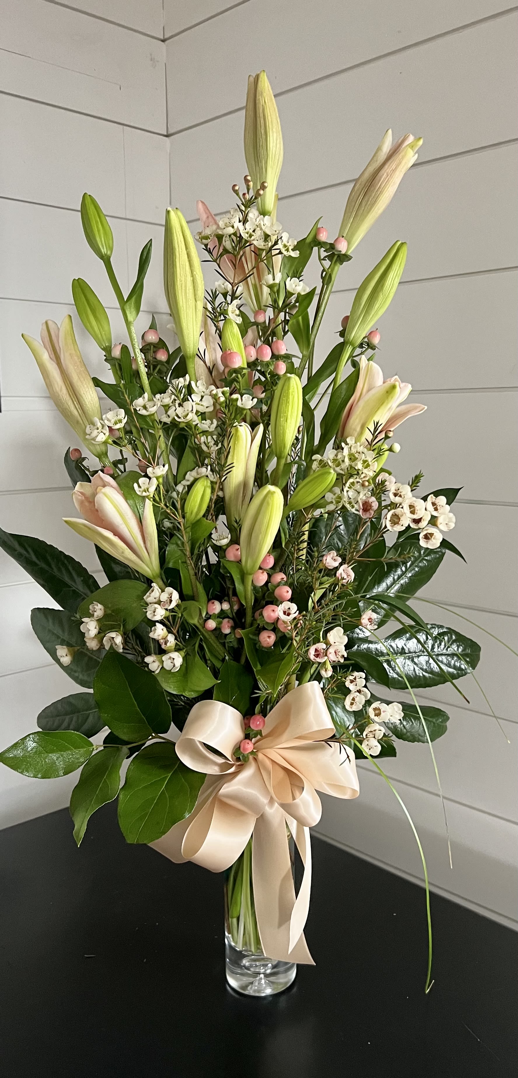 Tall bouquet of pale lilies and white filler flowers in a glass vase with a satin bow