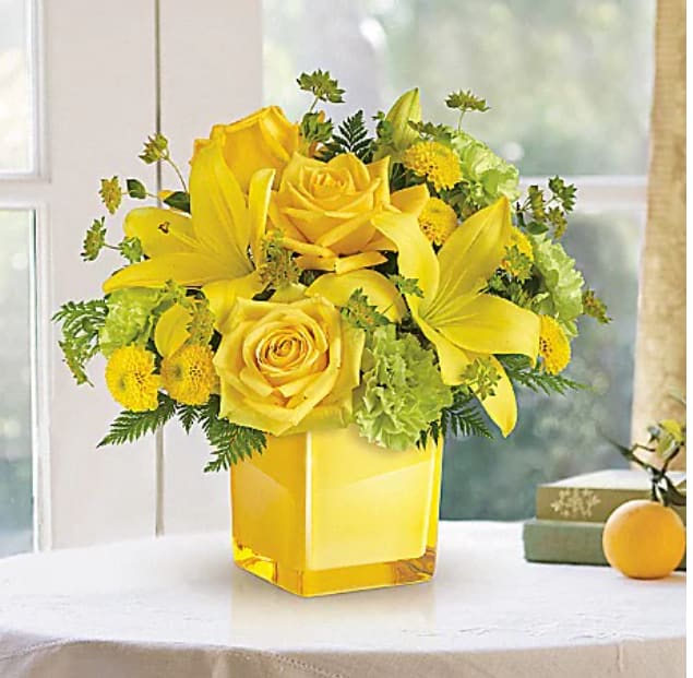 Sunny Mood Bouquet - An instant pick-me-up! Beautifully arranged in a stylish cube vase, these radiant roses and lilies deliver smiles and sunshine, any day of the week. This bright bouquet includes yellow roses, yellow asiatic lilies, green carnations, yellow button spray chrysanthemums, bupleurum and leatherleaf fern. Delivered in a glass cube. 