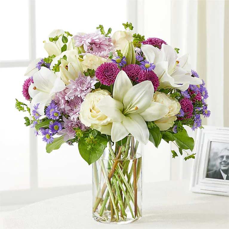 Bouquet of white lilies, cream roses, and purple flowers in a clear glass vase
