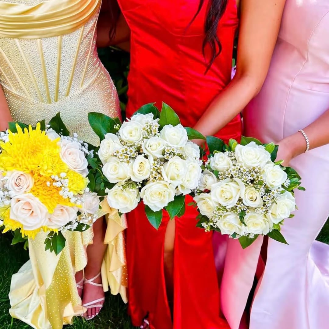 Three women hold bouquets of white and yellow roses in formal dresses.