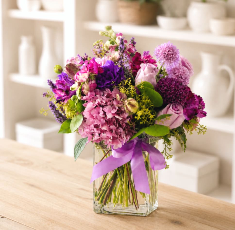 Mixed pink and purple bouquet in a clear square vase with a lavender ribbon