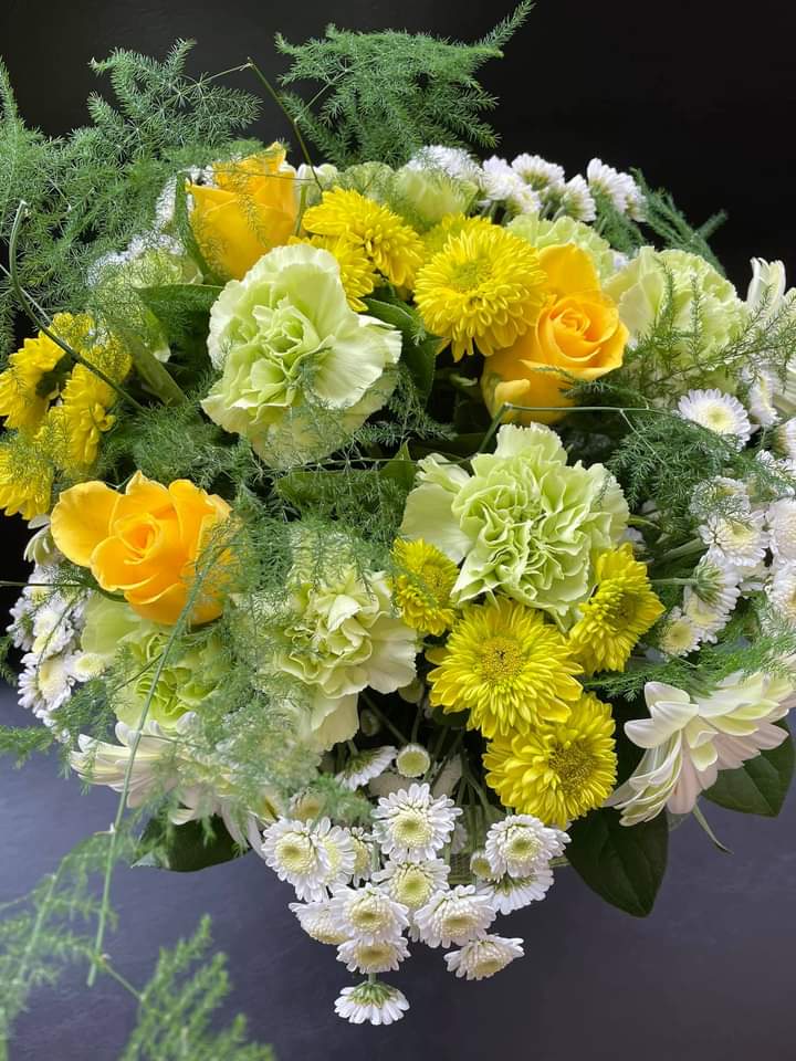 Round arrangement of yellow roses, green carnations, white chrysanthemums, and airy fern-like greenery.