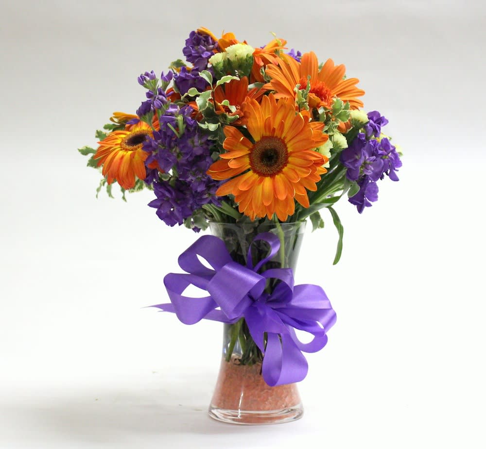 Orange and purple mixed bouquet in a clear glass vase with a purple ribbon