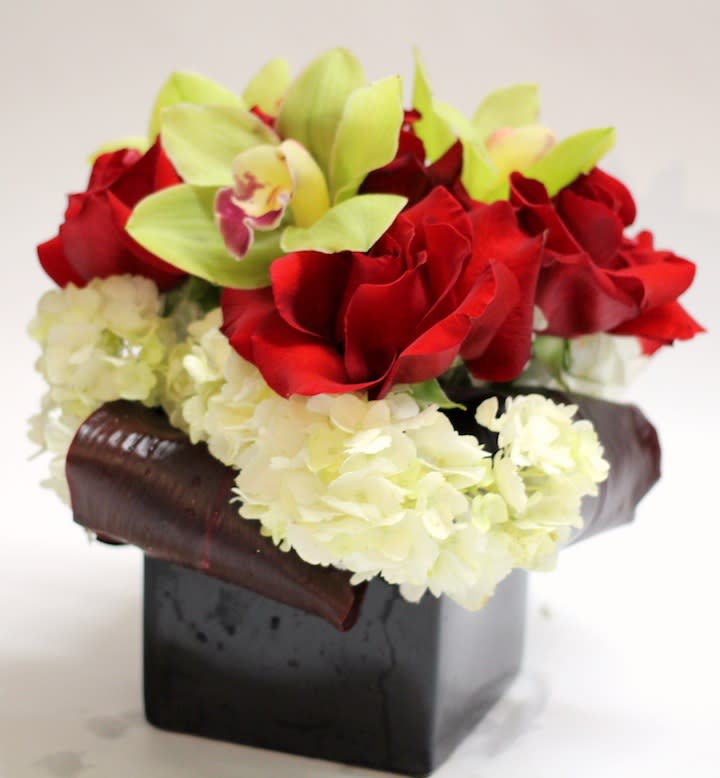 Red roses and pale green orchids in a black square vase with white hydrangeas