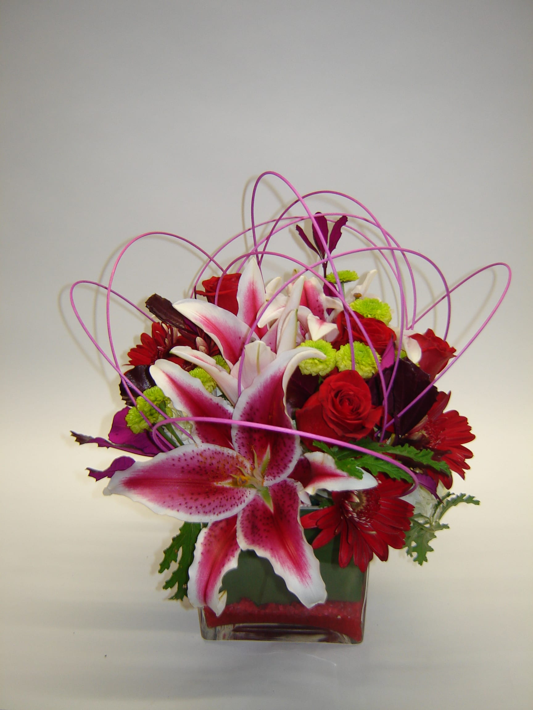 Pink lilies and red roses in a square glass vase with looping purple accents