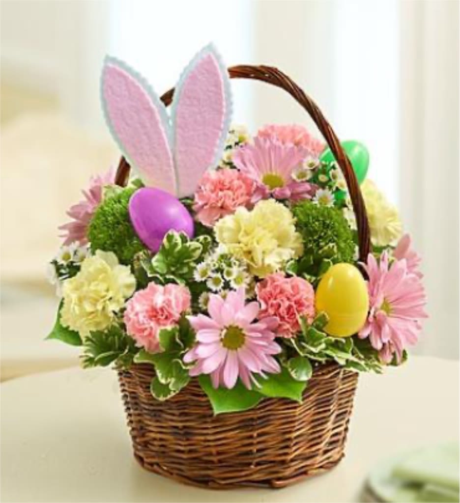 Springtime Bunny Bliss - This charming arrangement captures the joyful spirit of spring with a playful Easter twist. Nestled in a rustic woven basket, soft pink daisies and delicate carnations bloom alongside fresh green accents, creating a gentle, pastel harmony. Bright decorative eggs peek through the blossoms, while adorable bunny ears rise above the bouquet, adding a whimsical touch. Perfect for celebrating new beginnings, this bouquet brings warmth, cheer, and a hint of magic to any space.