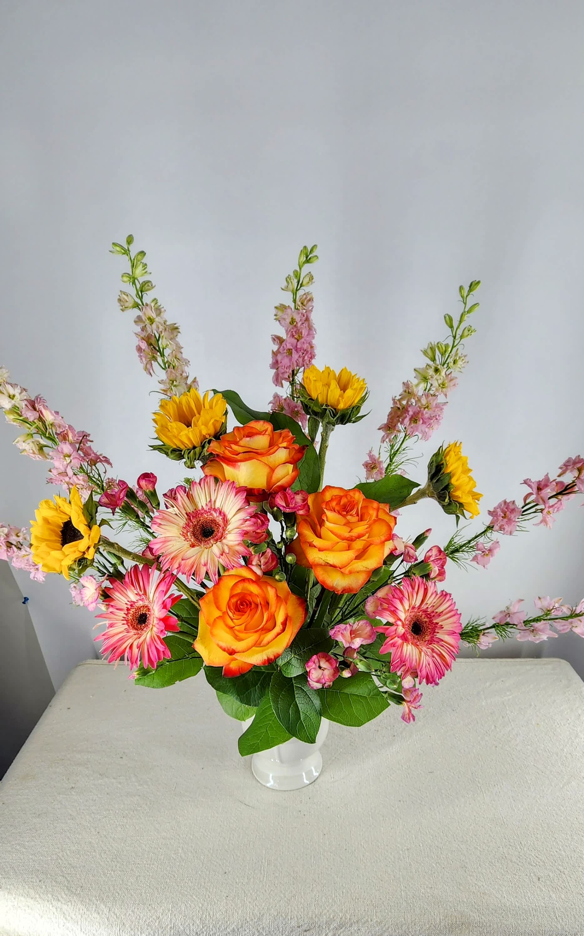Mixed bouquet of orange roses, pink gerberas, and yellow sunflowers in a white vase