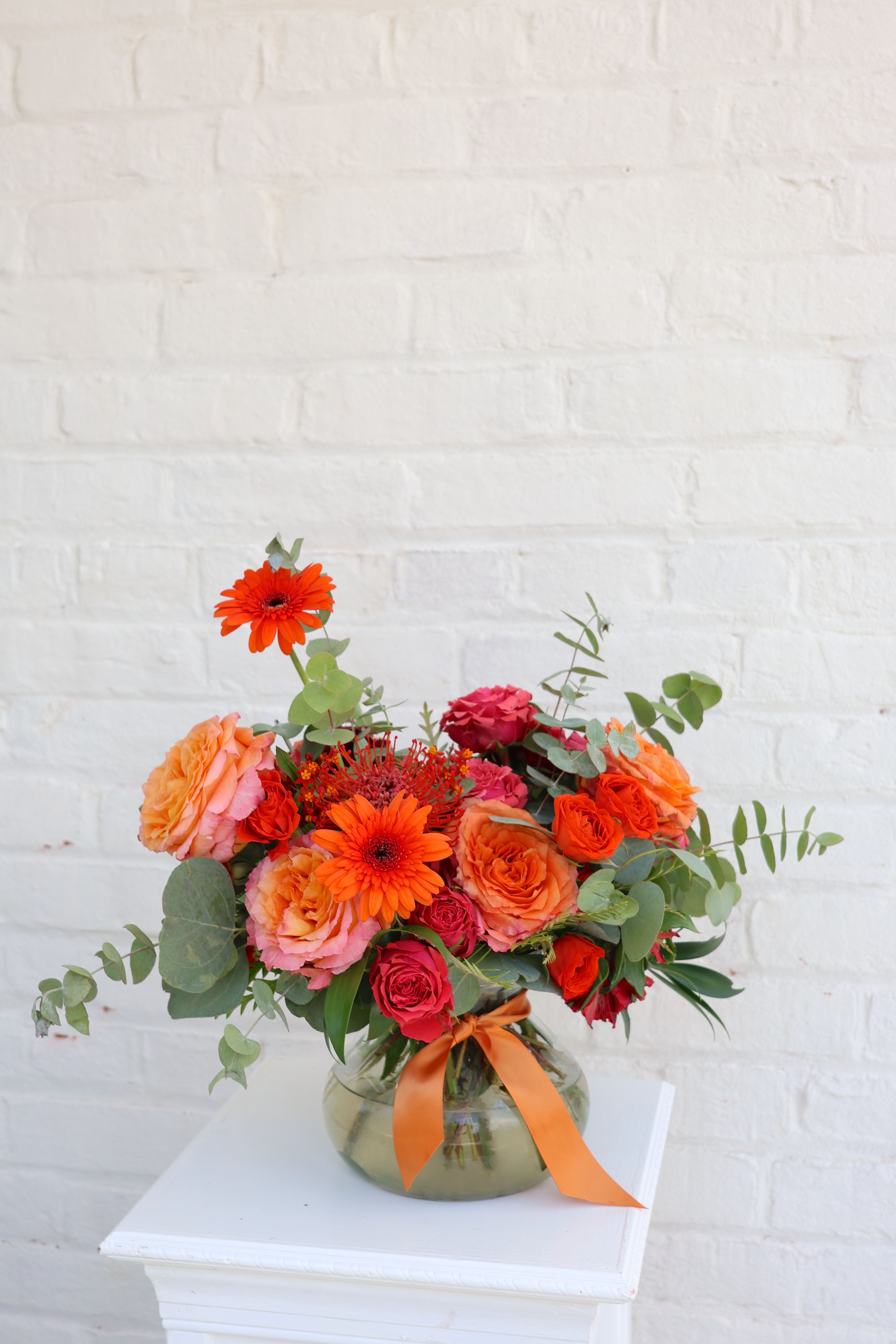 Orange and pink mixed flower arrangement in a glass vase with ribbon