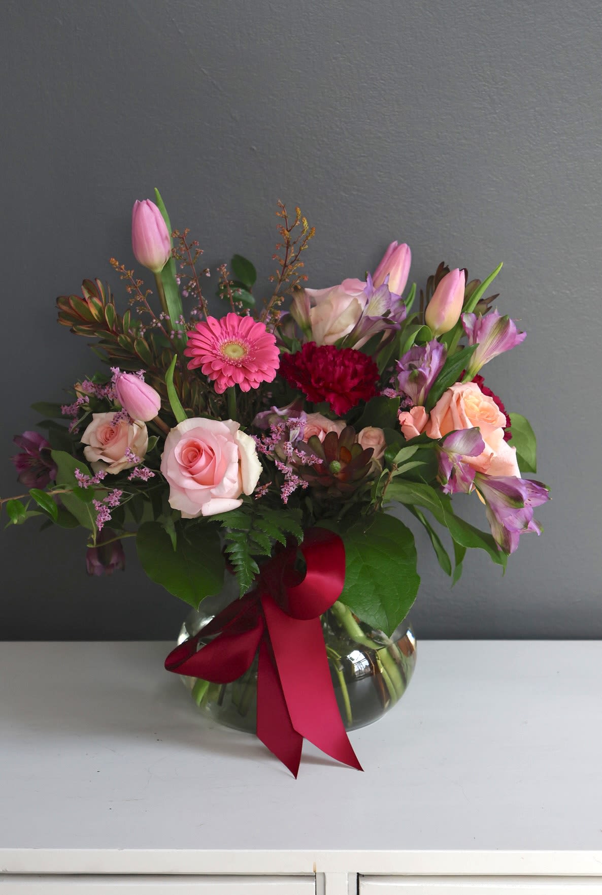 Pink roses and mixed blooms arranged in a glass vase with a red ribbon