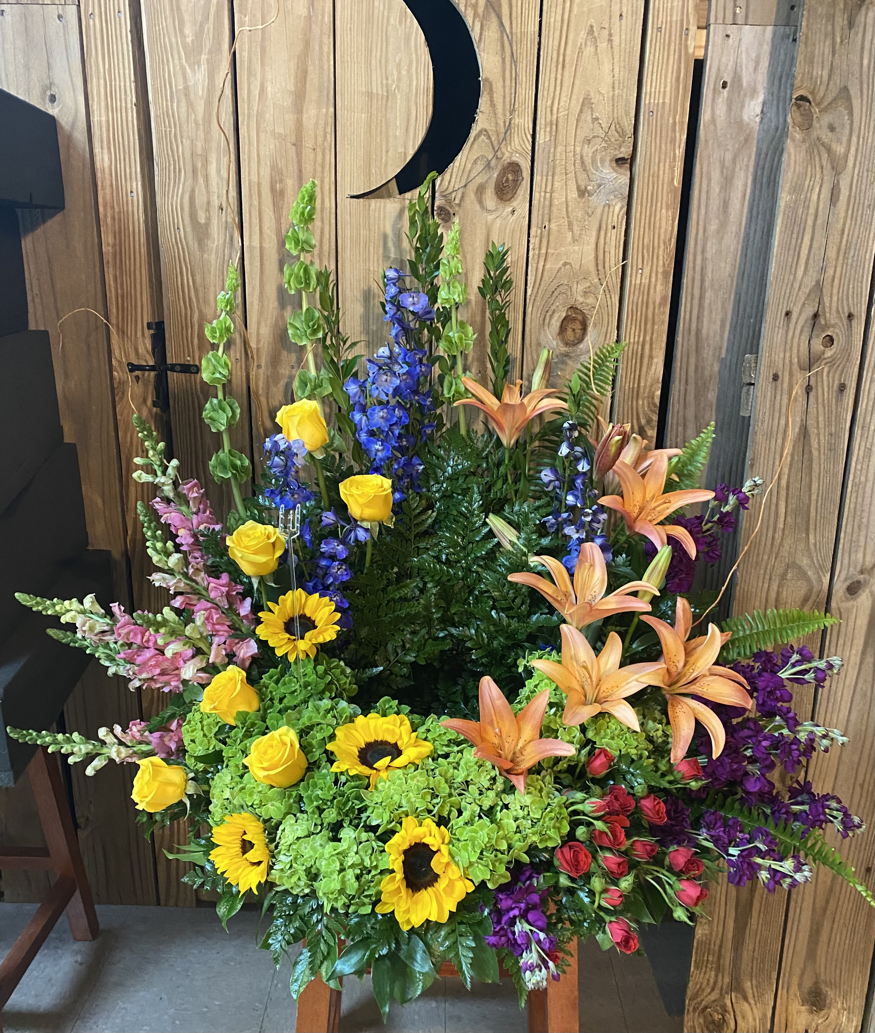 Tall floral arrangement with sunflowers, roses, lilies, and blue delphinium