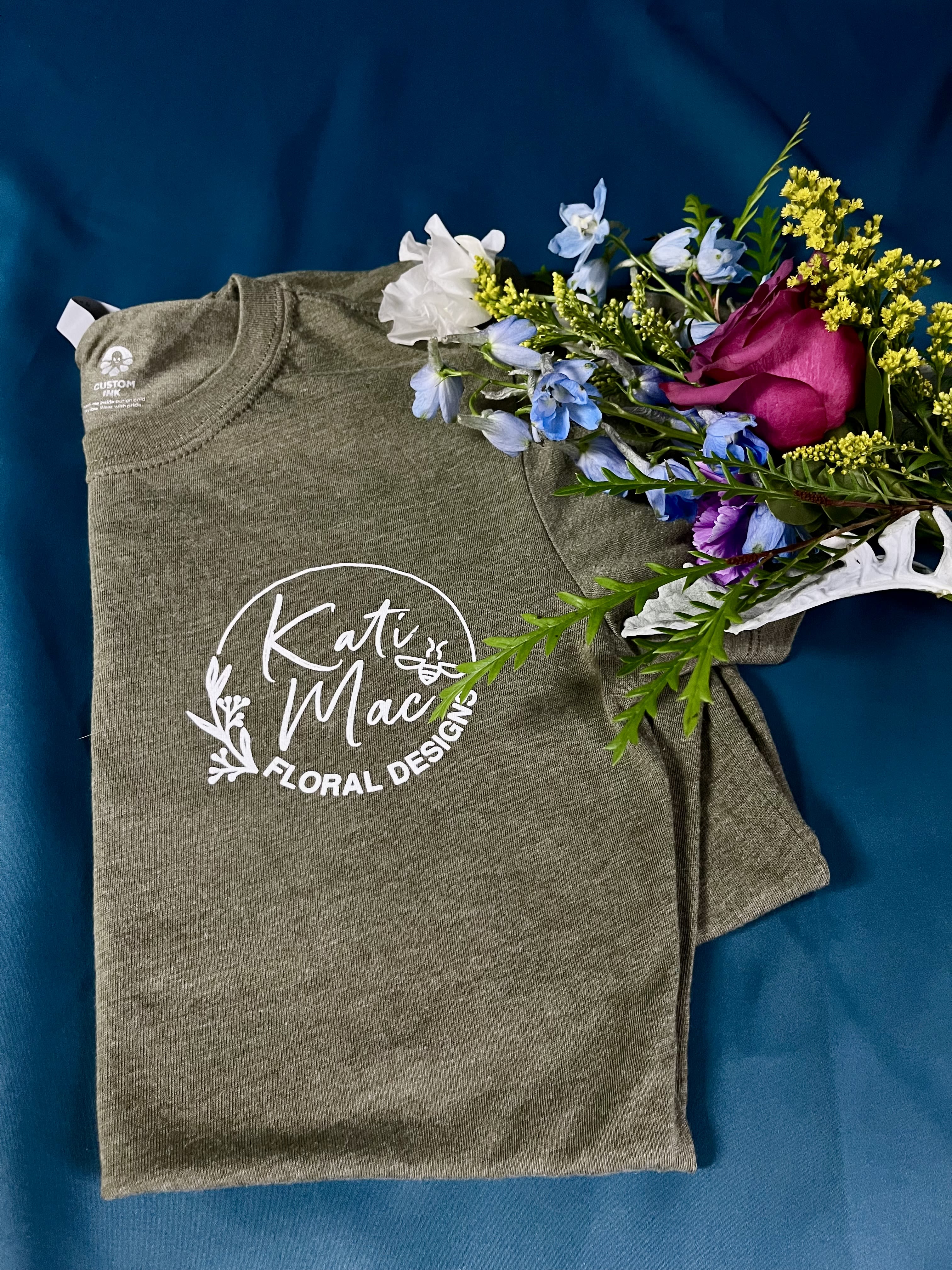 Folded green logo t-shirt with a small bouquet of pink rose and blue and yellow flowers on top