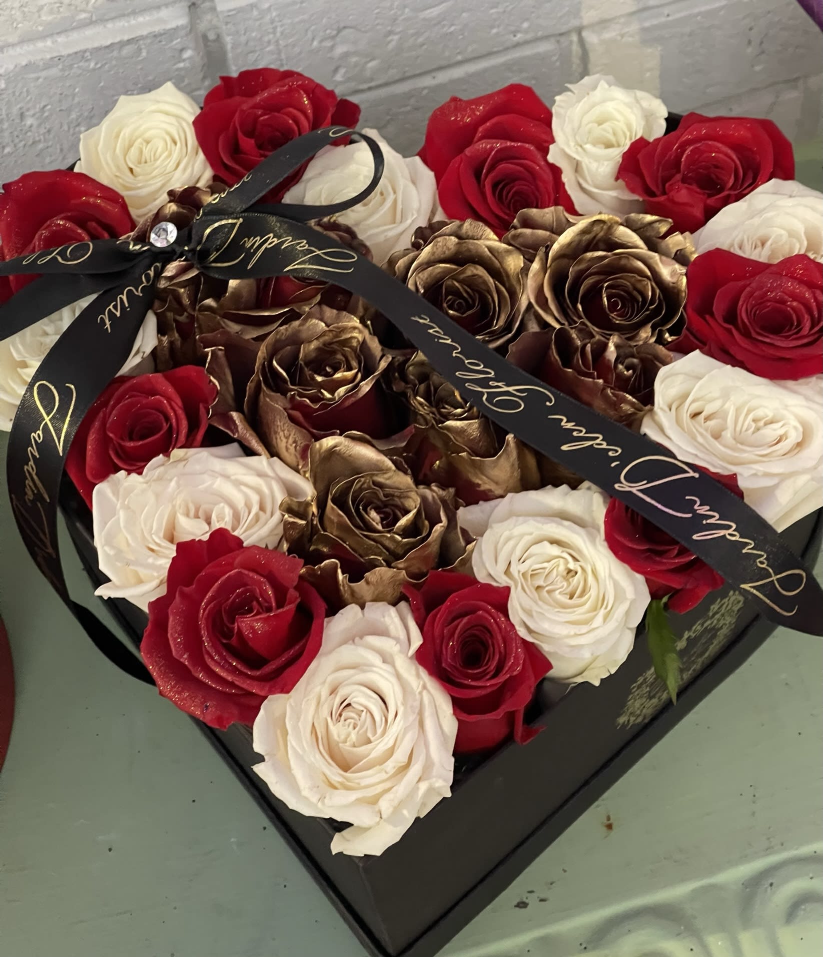 Heart-shaped box filled with red, white, and gold roses tied with a black ribbon