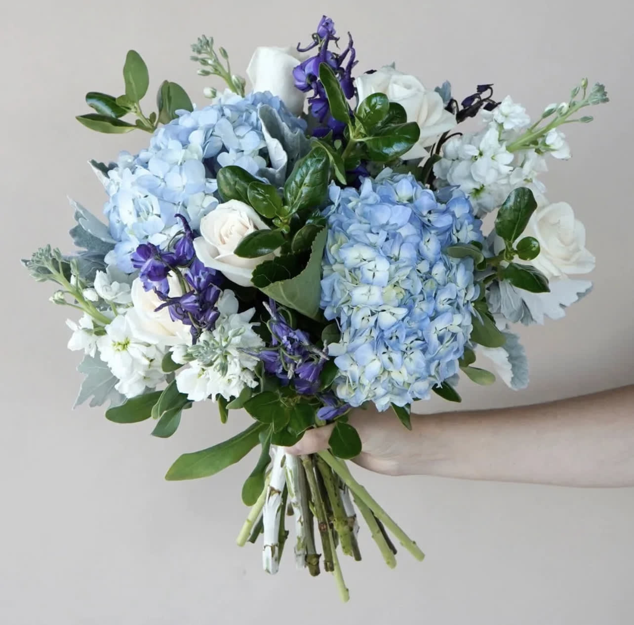 Handheld bouquet of blue hydrangeas, white roses, and purple flowers