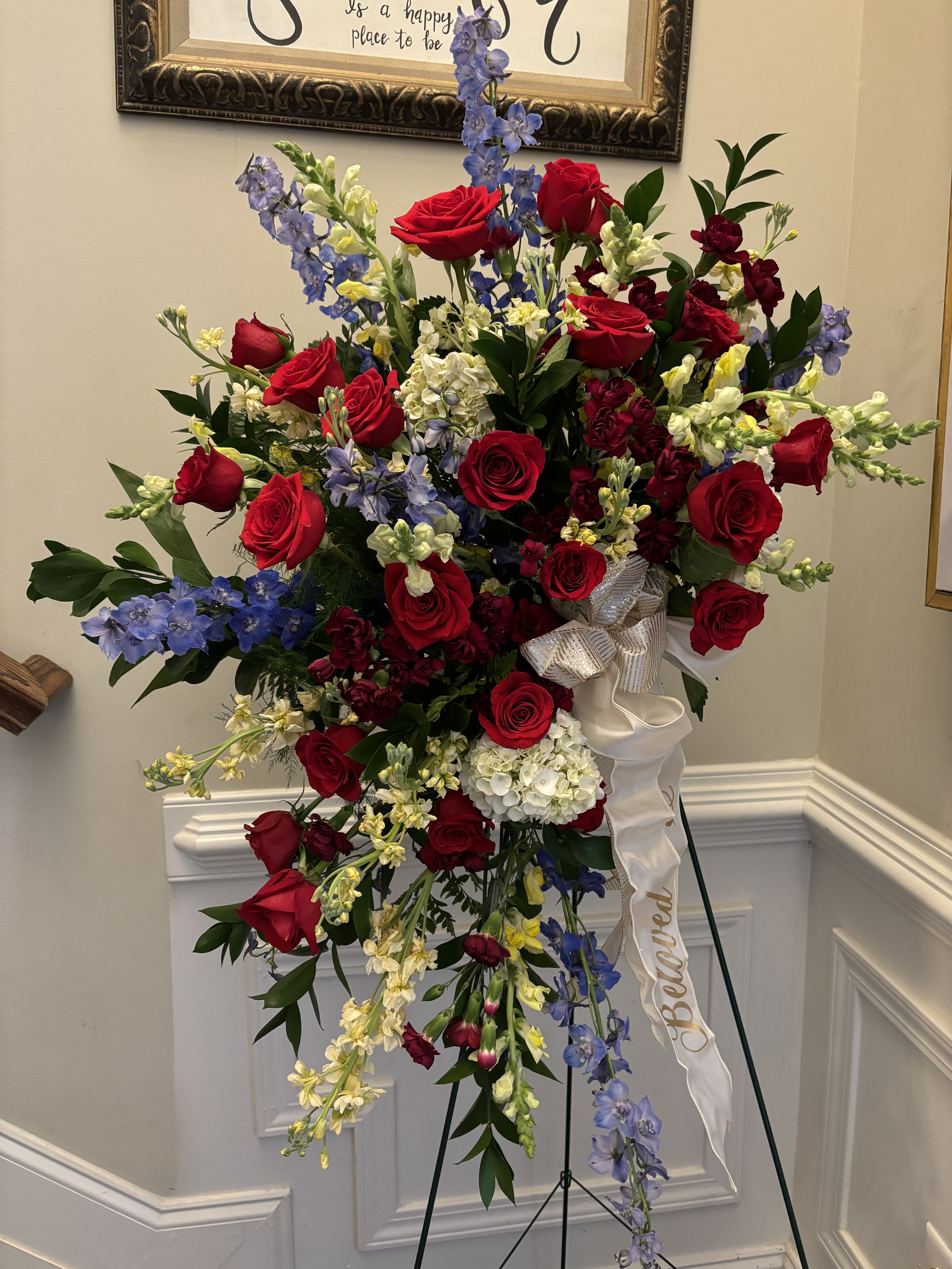 Broadway - Make the last gift to your loved one a flower standing spray to remember! With an arrangement  of roses snapdragons &amp; stock and a variety of seasonal greens placed around a stunning bow with the word “Beloved” detailed in a brush gold.   Arrangement can be made all white or in a variety of other colors.   Bow can also be specialized to say Mother, Father,etc 