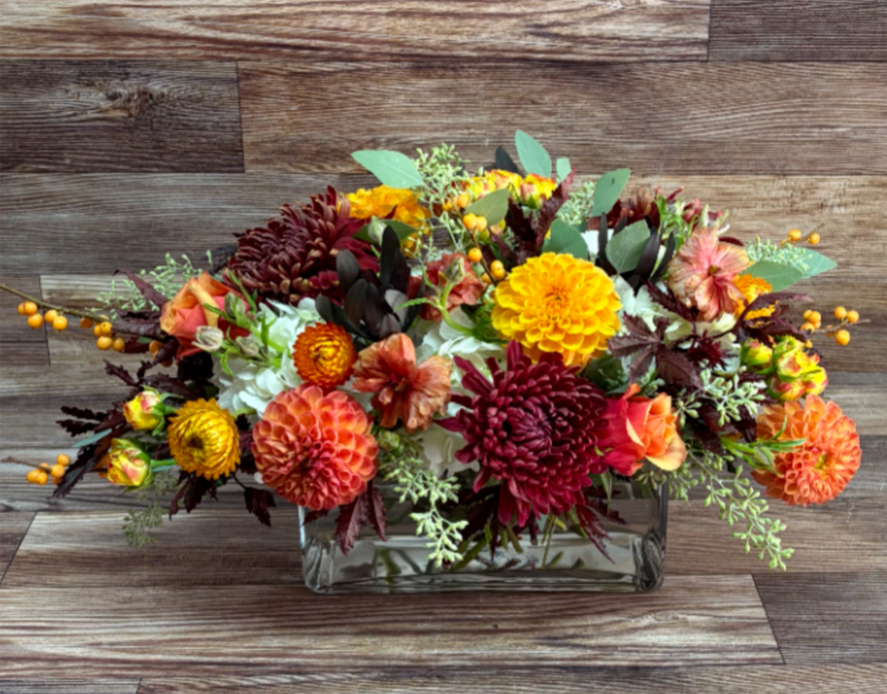 Low floral arrangement in a clear rectangular vase with orange, yellow, burgundy, and white blooms