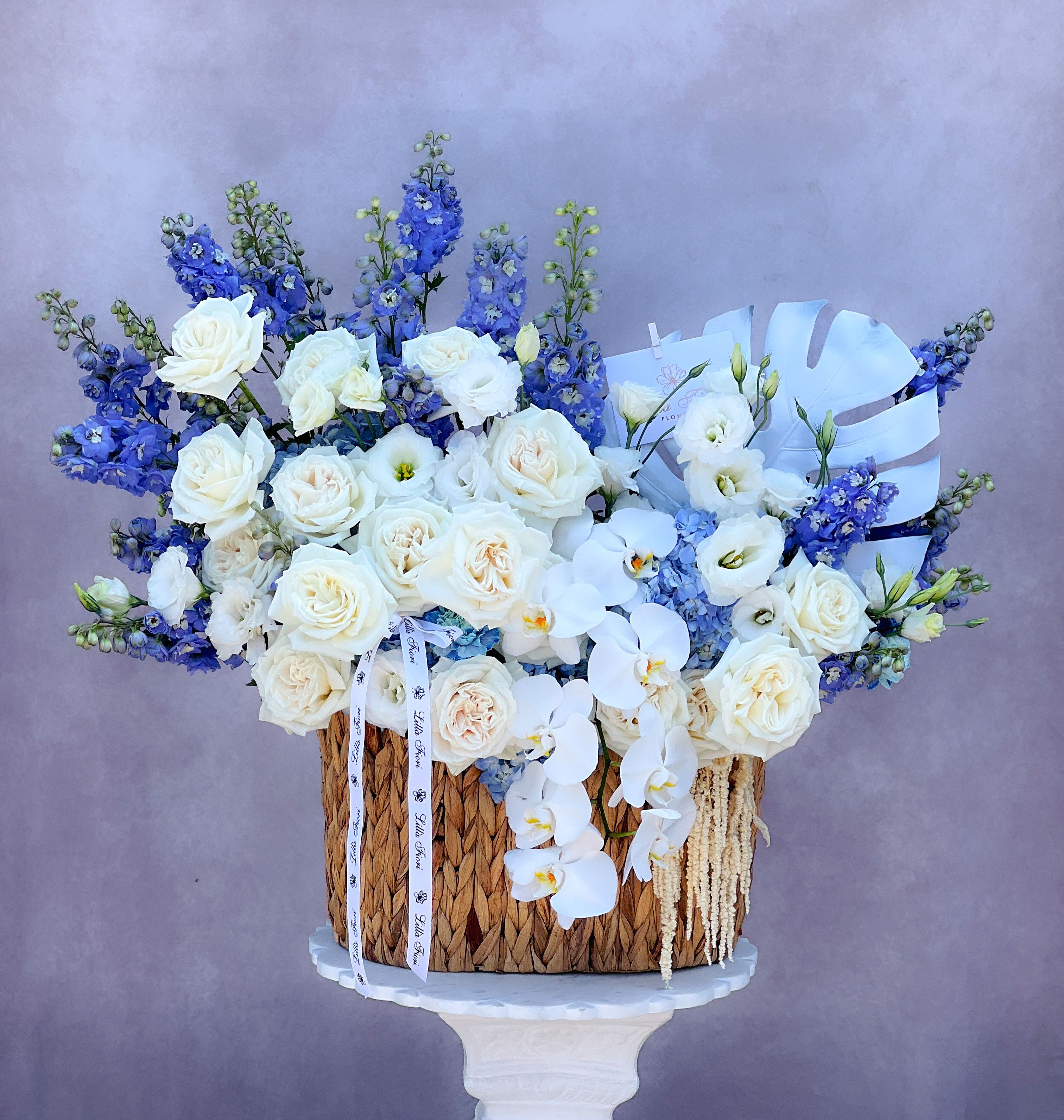 White roses and orchids arranged with blue flowers in a woven basket