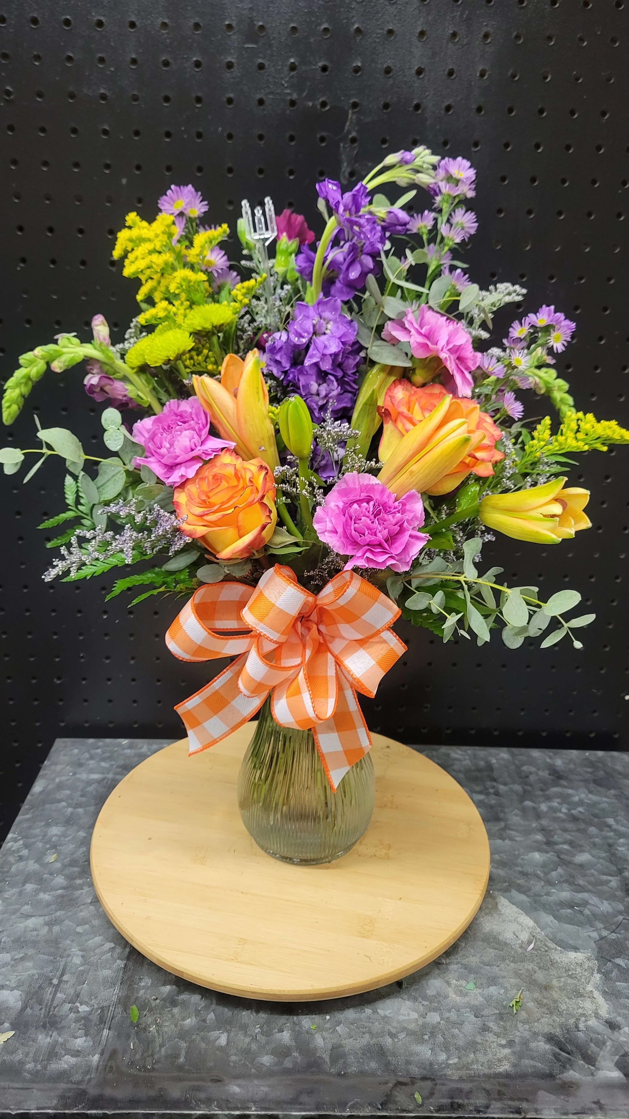 Mixed bouquet of orange roses, yellow lilies, and purple flowers in a glass vase