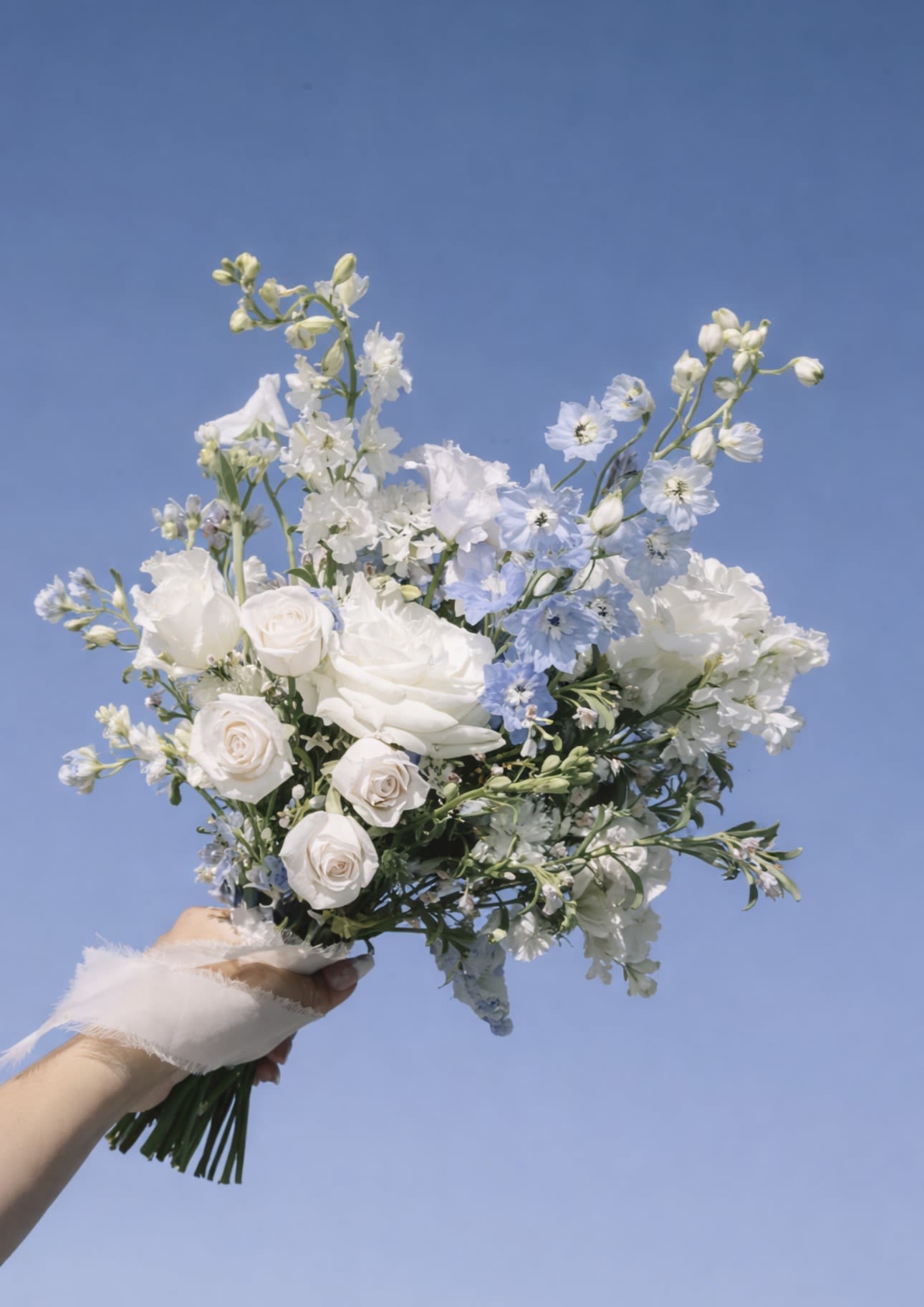 Handheld bouquet of white and pale blue flowers against a blue sky