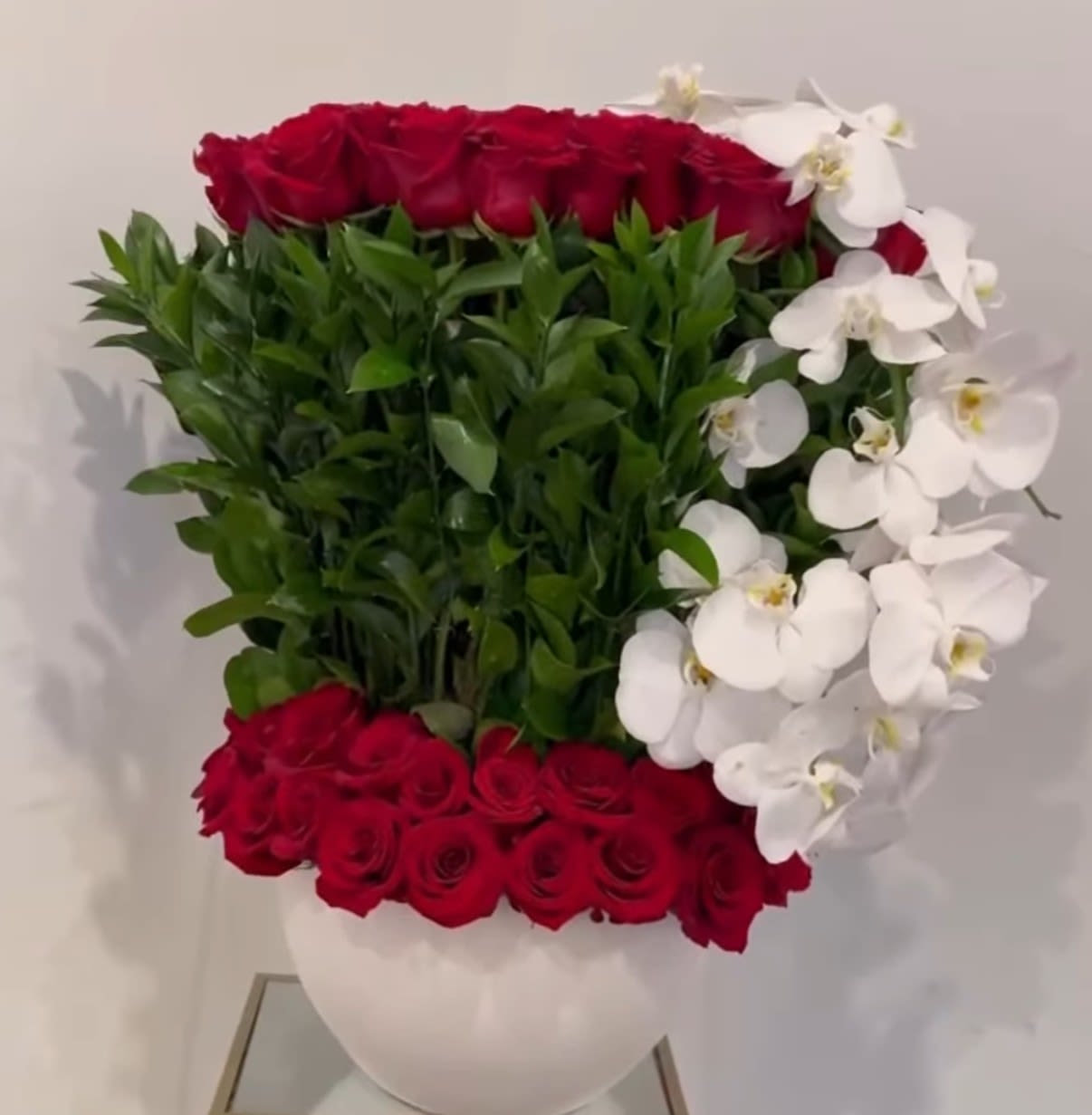 Red Velvet Orchid - An extraordinary floral statement designed to captivate. This luxurious arrangement features approximately 70 premium red roses, artfully combined with elegant white Phalaenopsis orchids, creating a stunning contrast of passion and sophistication.  Designed in a modern bottle-style vase, the composition is enhanced with lush ruscus greenery, adding texture and a refined, natural flow. Every element is carefully arranged to achieve a full, elevated look that feels both timeless and contemporary.  Perfect for grand romantic gestures, anniversaries, or unforgettable celebrations, this arrangement speaks of deep love, elegance, and exclusivity.  Includes:  ~70 premium red roses White Phalaenopsis orchids Fresh ruscus greenery Decorative bottle-style vase  A true luxury piece made to impress and leave a lasting impression. ️