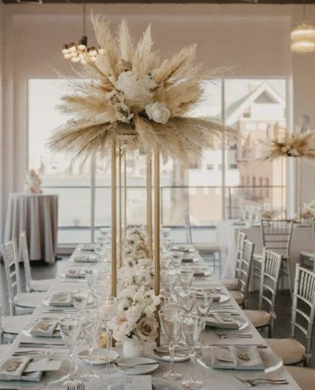 Elevated Boho Luxe Centerpiece - Bring sophistication and modern elegance to your reception with our Elevated Boho Luxe Centerpiece. Designed to create visual impact without obstructing guests' conversation, this tall centerpiece features a sleek gold metal stand topped with an abundant arrangement of preserved pampas grass, dried botanicals, ivory roses, and premium white florals. A coordinating floral accent at the base adds fullness and balance, creating a luxurious tablescape perfect for upscale weddings.  Ideal for bohemian, modern, garden, and luxury celebrations, this centerpiece transforms your reception into an unforgettable experience.  Includes:  Tall gold metal centerpiece stand Premium preserved pampas grass Dried botanical accents Ivory roses and premium white florals Coordinating floral arrangement at the base Professional floral design and styling