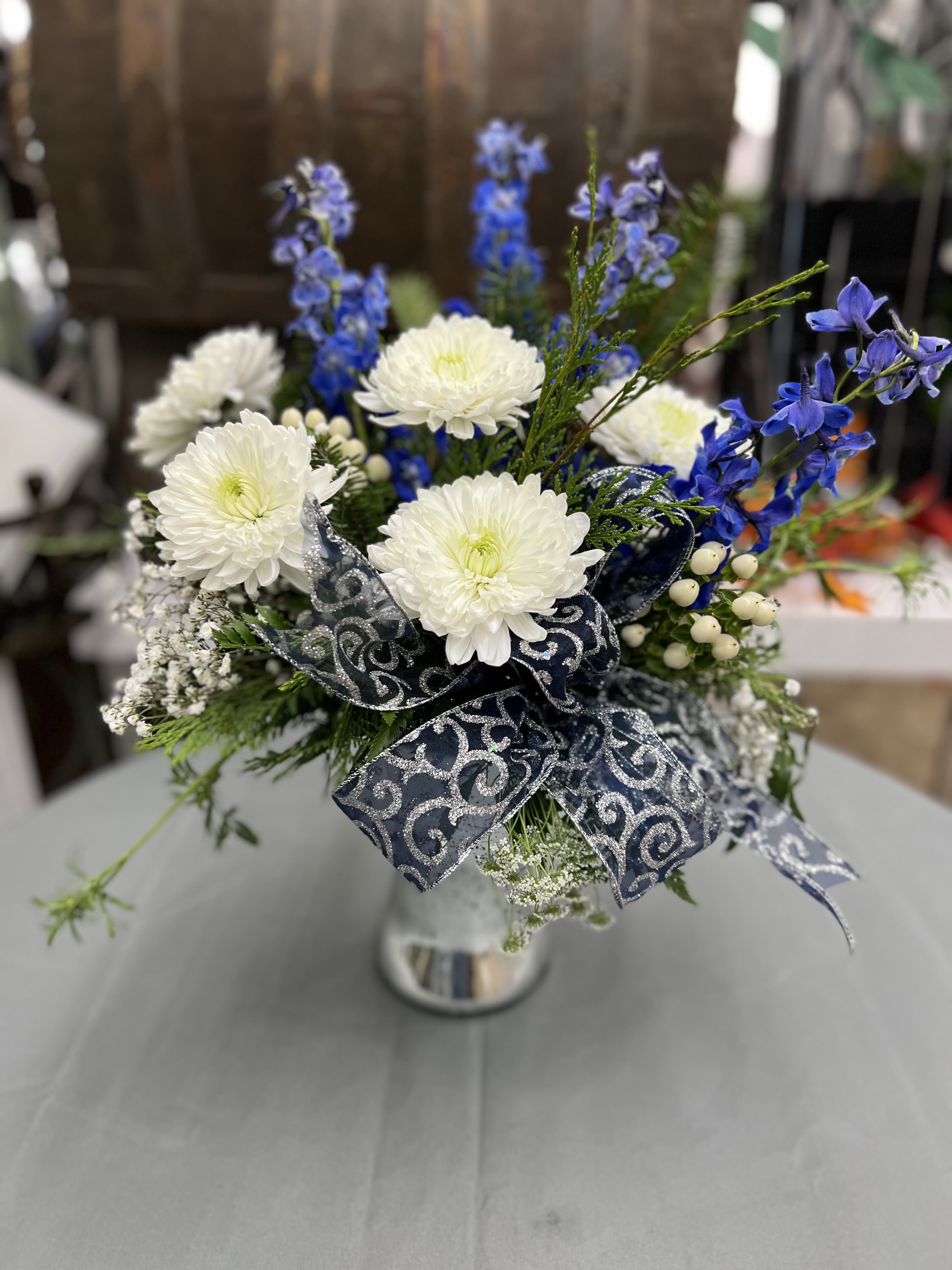 White chrysanthemums and blue flowers in a silver vase with a patterned ribbon