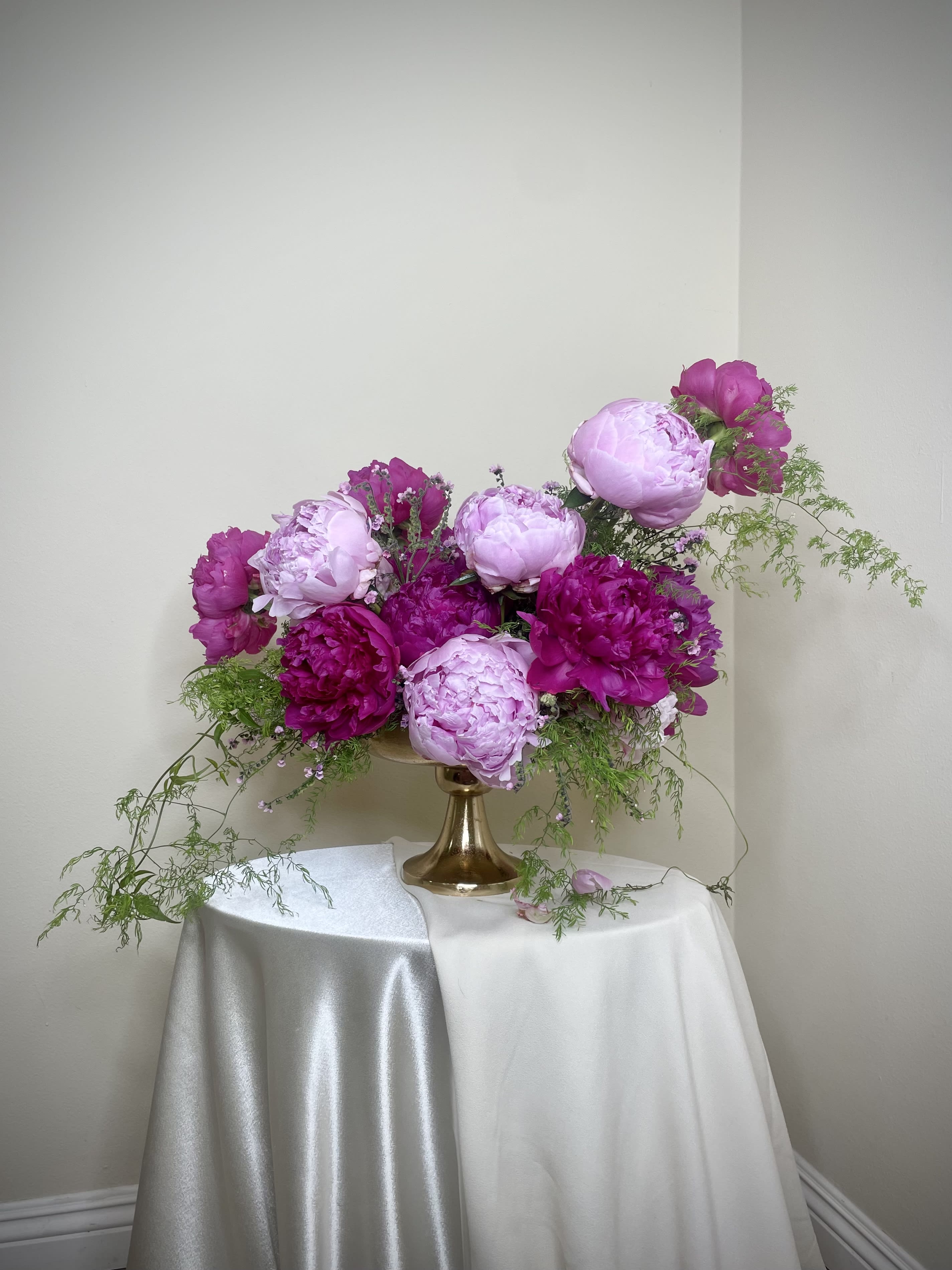 Low compote arrangement of pink and magenta peonies with trailing greenery in a gold pedestal vase.