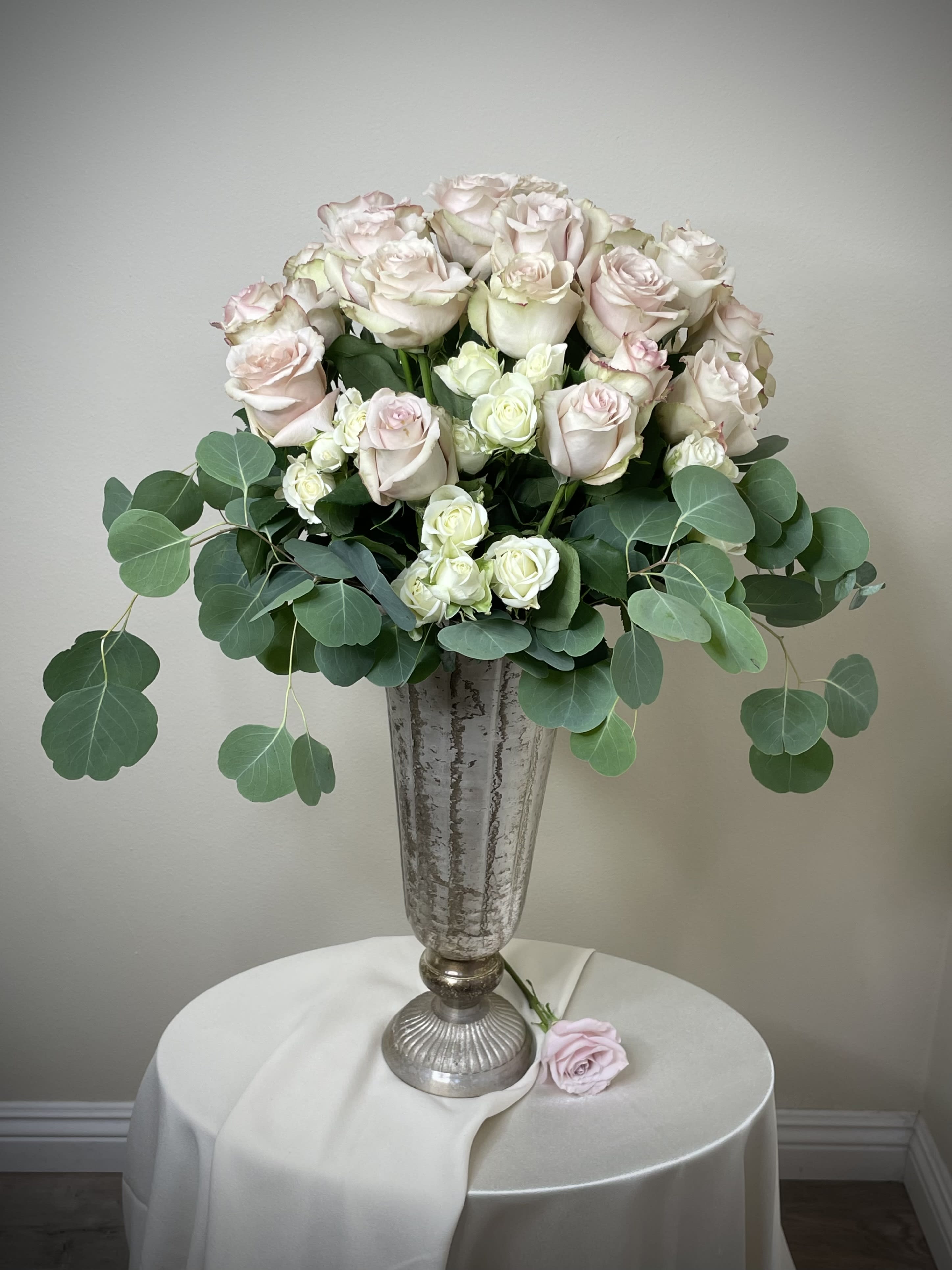 Tall silver urn with blush and ivory roses and greenery on a draped round table