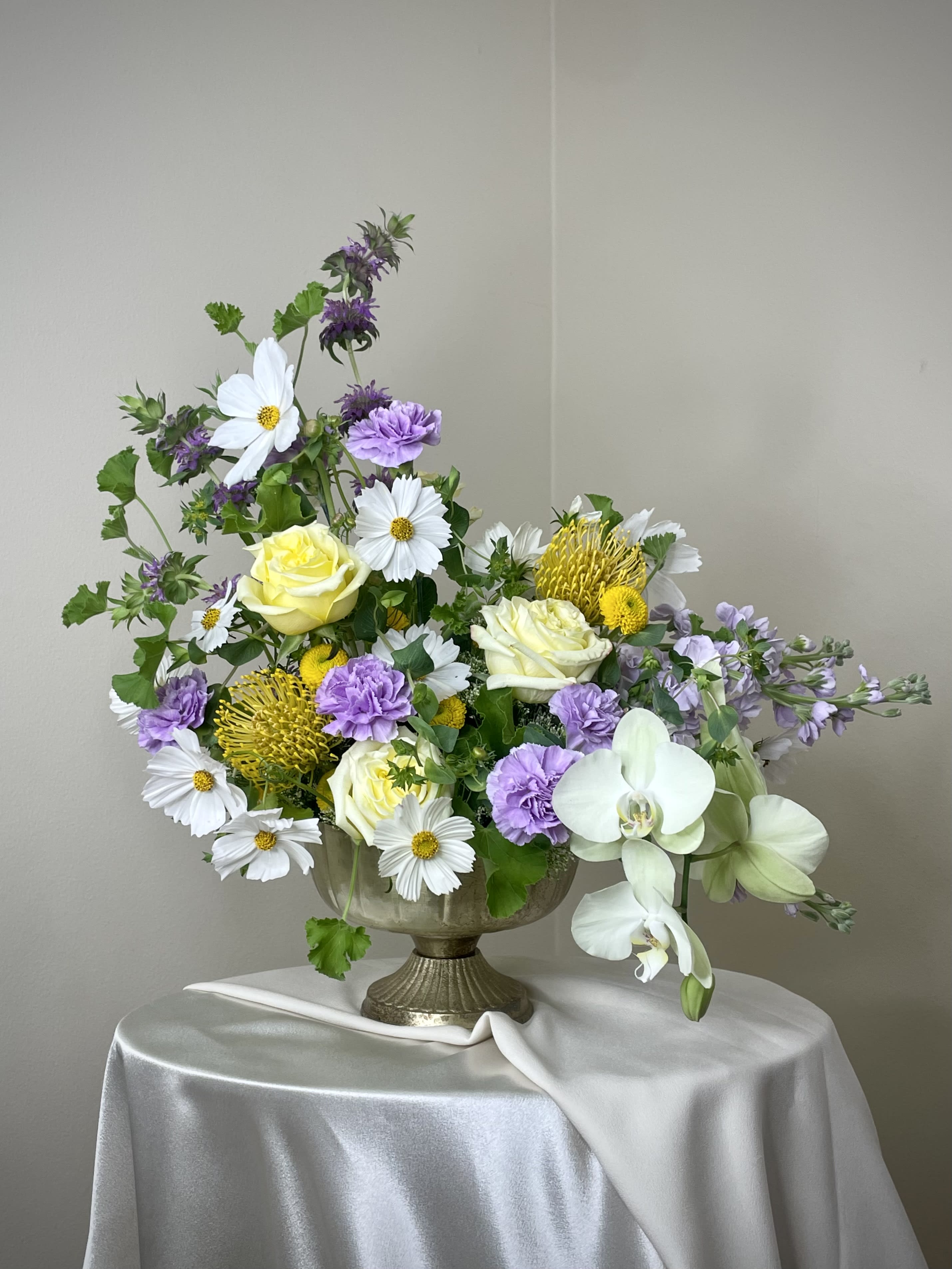 Garden-style arrangement of white, yellow, and lavender flowers in a metal pedestal bowl on a draped table.