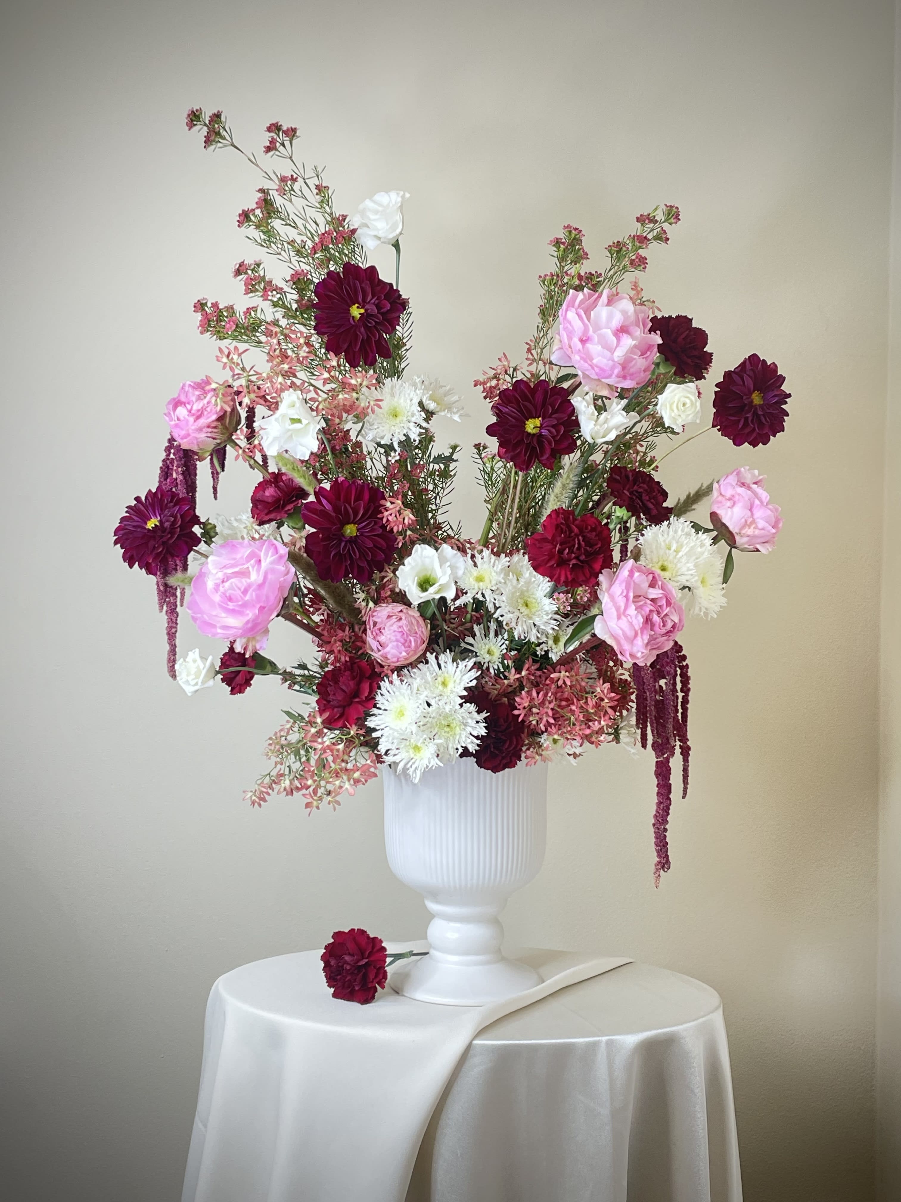 Tall arrangement of burgundy, pink, and white flowers in a white urn on a draped round table