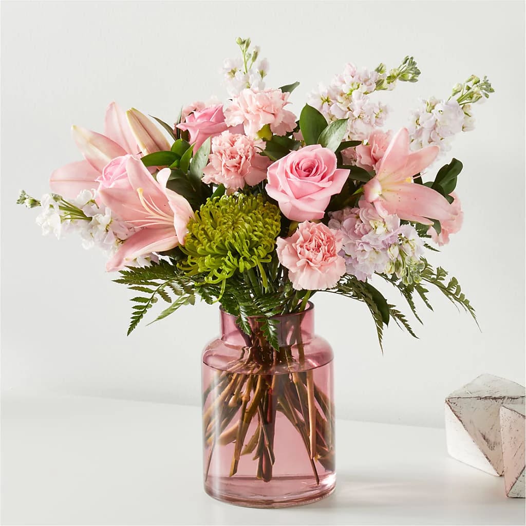 Pink Bouquet - Make their day with this delicately gorgeous, softly hued bouquet of pink roses, lilies, carnations, Fuji mums, Israeli ruscus, and leatherleaf in a matching blush vase.  Please Note: The bouquet pictured reflects our original design for this product. While we always try to follow the color palette, we may replace stems to deliver the freshest bouquet possible, and we may sometimes need to use a different vase. DETAILS Designed by florists, ready to display. For long–lasting blooms, replace the water daily. We suggest trimming the stems every couple days. Pet Safety Precautions: This bouquet or plant may include flowers and foliage that are known to be toxic to pets. To keep them safe, be sure to keep this arrangement out of your pet's reach.