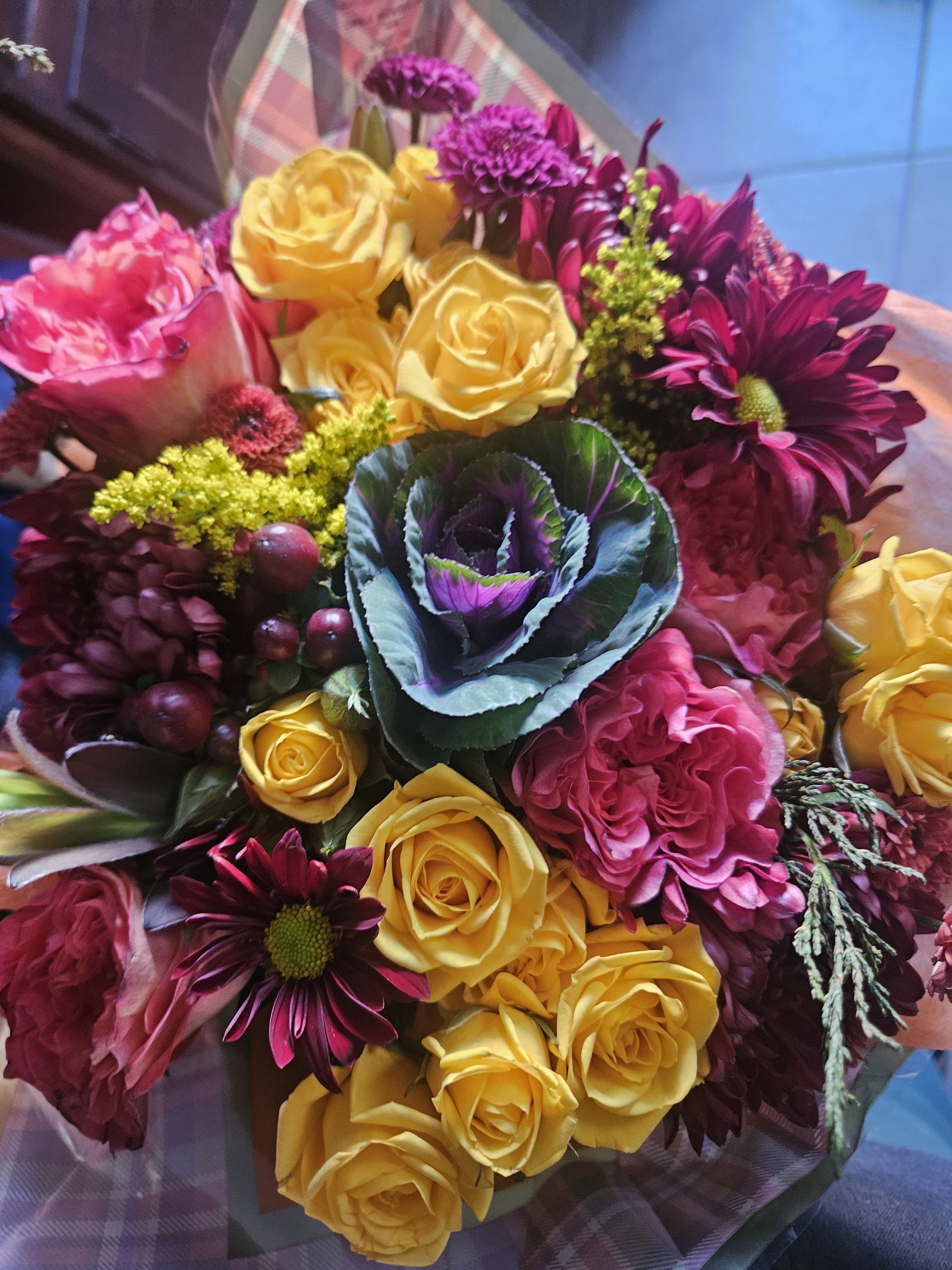 Hand-tied bouquet of yellow roses, pink blooms, purple mums, and ornamental cabbage in plaid wrap