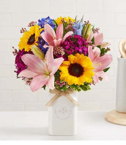 Honey Bee Buzz - Get them buzzing with our charming bouquet! Sunflowers, Pink Asiatic Lily, Blue Delphinium, Fuchsia Dianthus, Pink Waxflower and Assorted Greens are gathered together for a mix of color and texture. Designed in our Sweet As Can Bee artisanal vase, featuring an adorable honey bee and raffia bow, it’s a gift that adds quaint beauty to any decor.