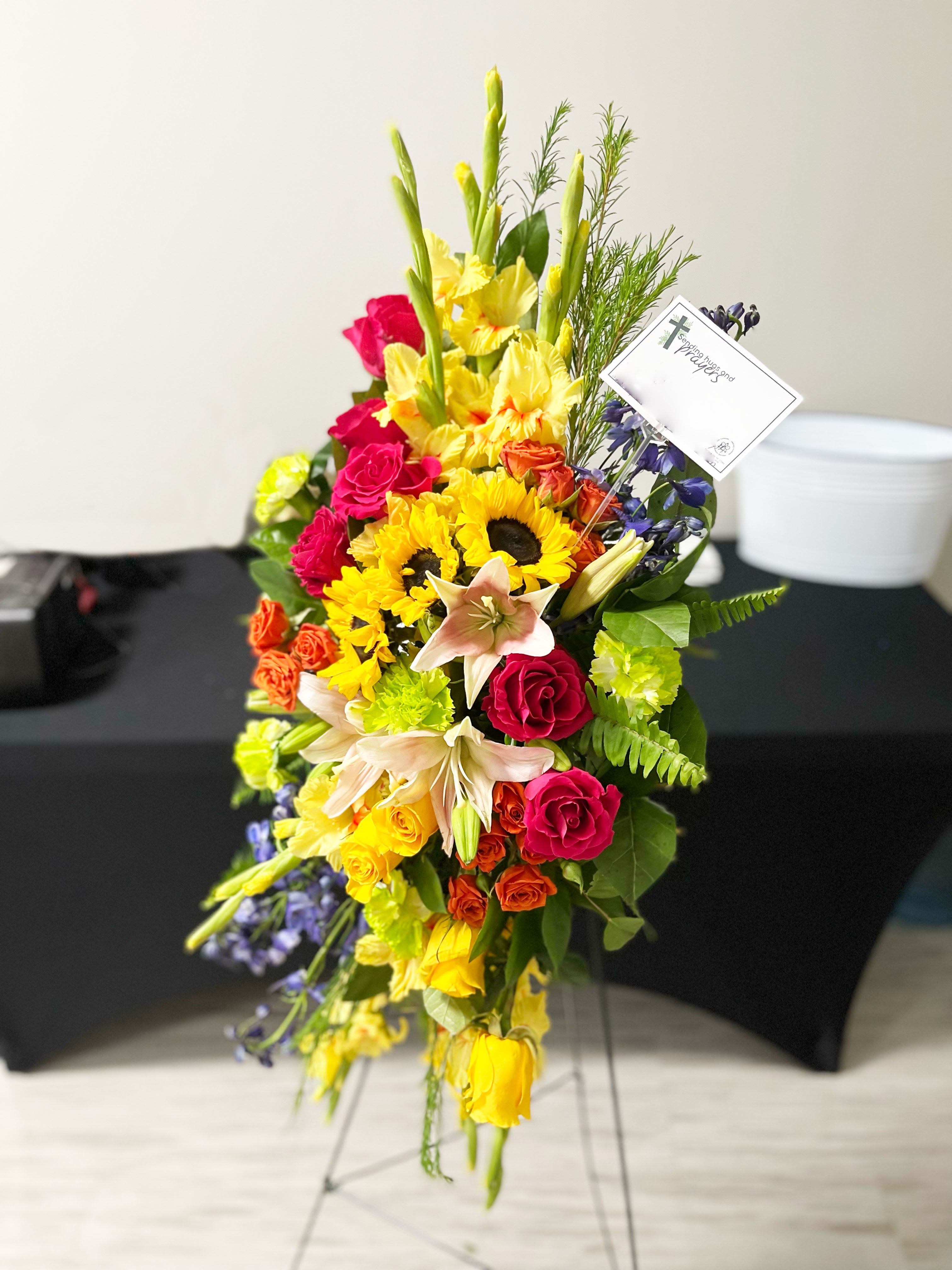 LOVE LIVES ON SPRAY - A bold, bright tribute, this rainbow of blooms fills the room with a spirit of hope and joy. What a glorious celebration of a love that will live on forever.  This spray features hot pink roses, yellow roses, orange spray roses, pink asiatic lilies, green gladioli, green carnations, medium yellow sunflowers, blue delphinium, bupleurum, myrtle, sword fern, silver dollar eucalyptus, and lemon leaf. Delivered on a wire easel.