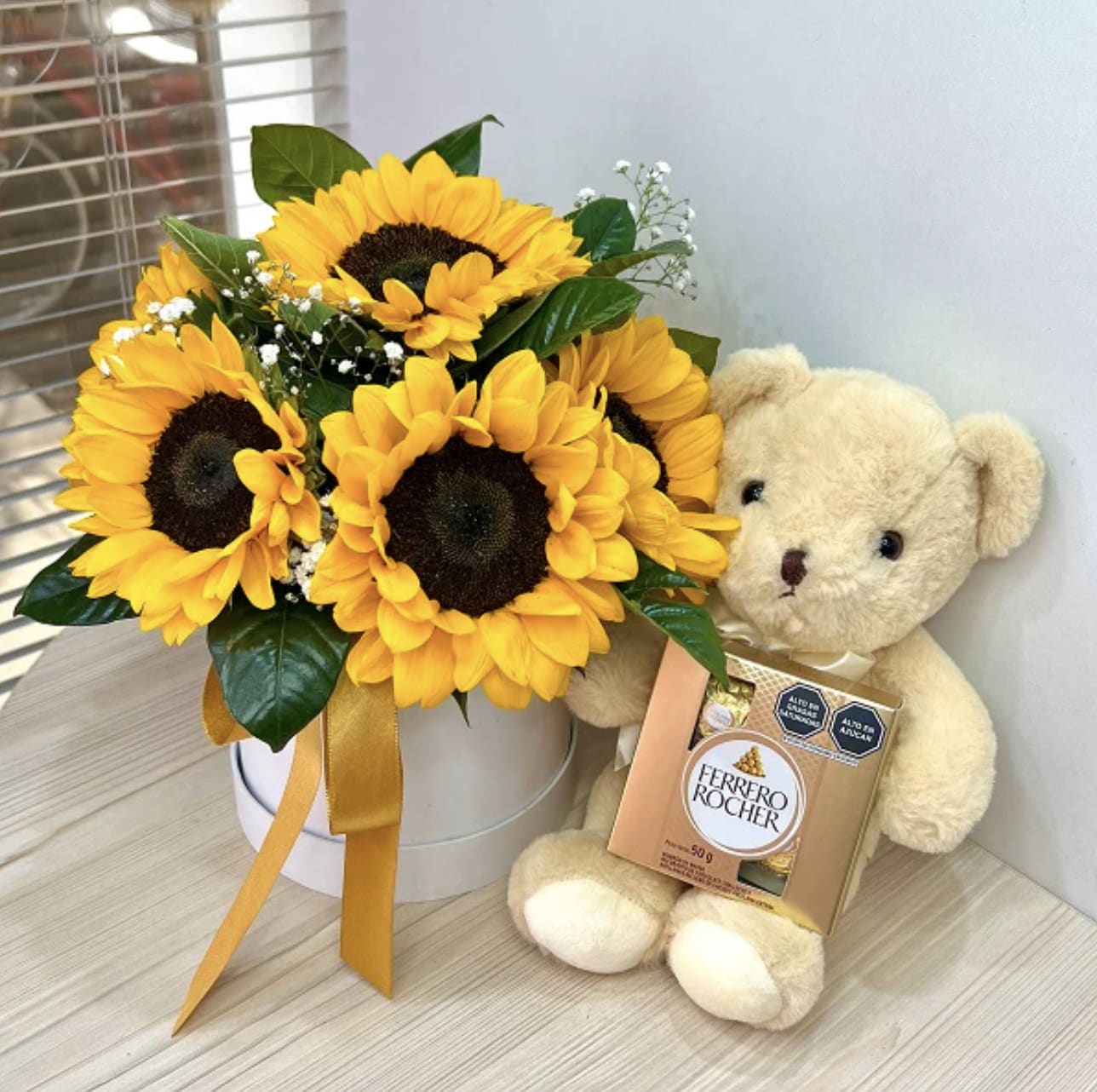 Box of Sunflowers - Surprise your loved ones with this delightful gift set, featuring a vibrant bouquet of fresh sunflowers beautifully arranged in a chic white hatbox and accented with lush green foliage and delicate baby’s breath. The cheerful flowers are perfectly complemented by an adorable small plush teddy bear (Note: Bear color will vary) holding a box of indulgent Ferrero Rocher chocolates. This thoughtful combination of blooms, sweetness, and cuddles is perfect for birthdays, anniversaries, or simply to spread joy.
